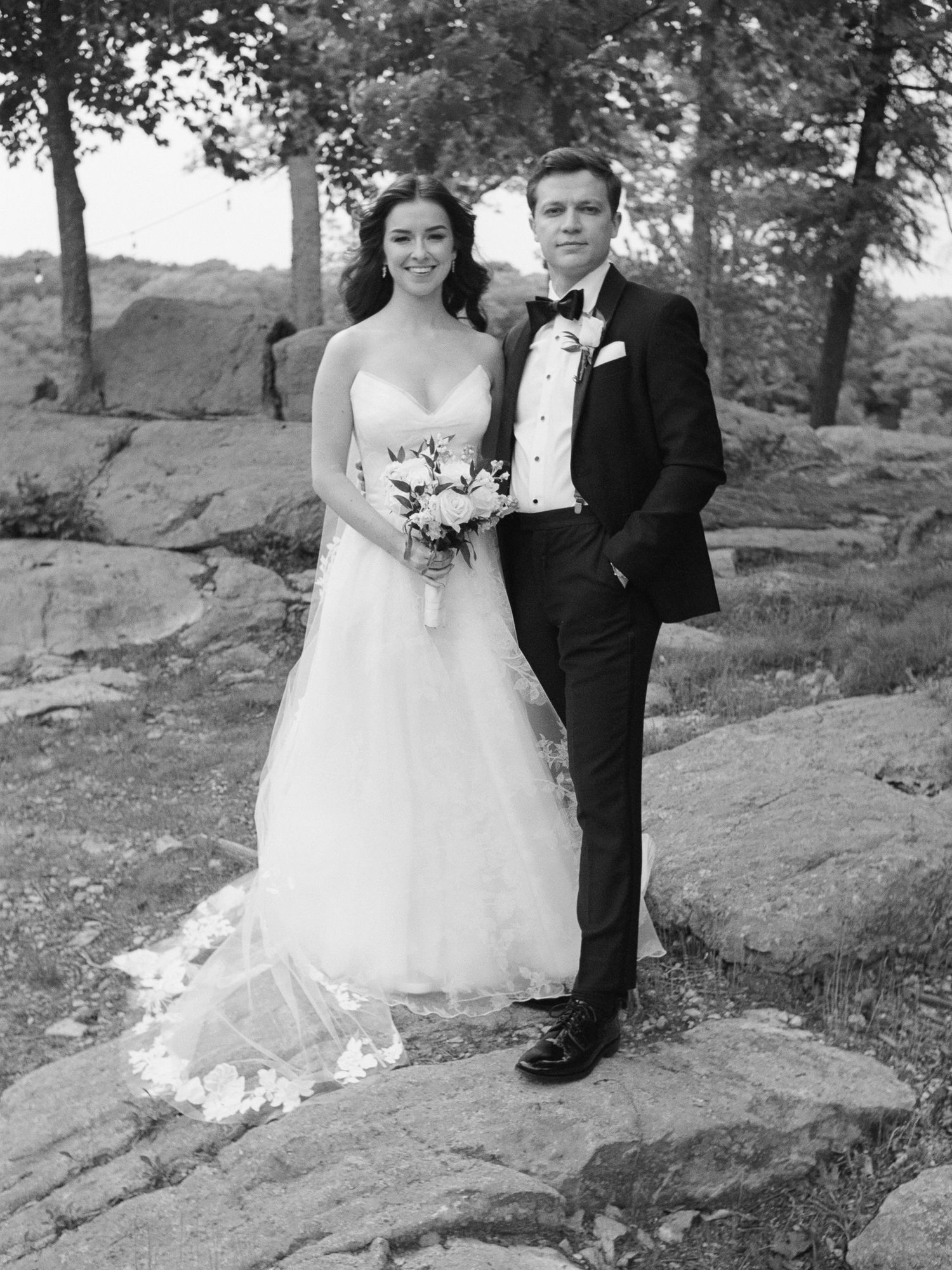 Bride and groom pose on rocky outcrop in black and white wedding portrait, she holds bouquet in elegant gown.