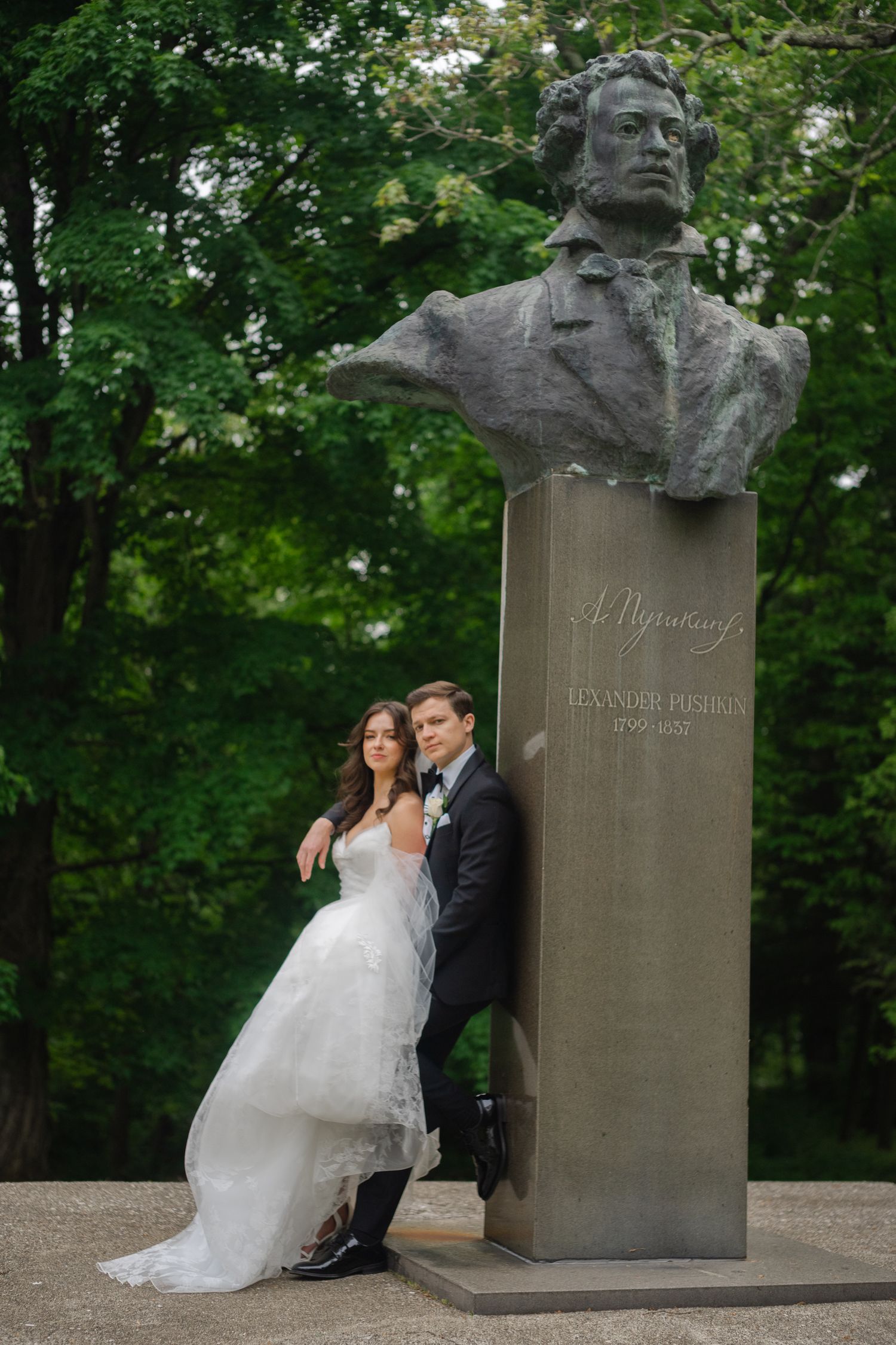 Wedding couple posing by Alexander Pushkin bust monument in lush green park, bride in white gown, groom in tuxedo.