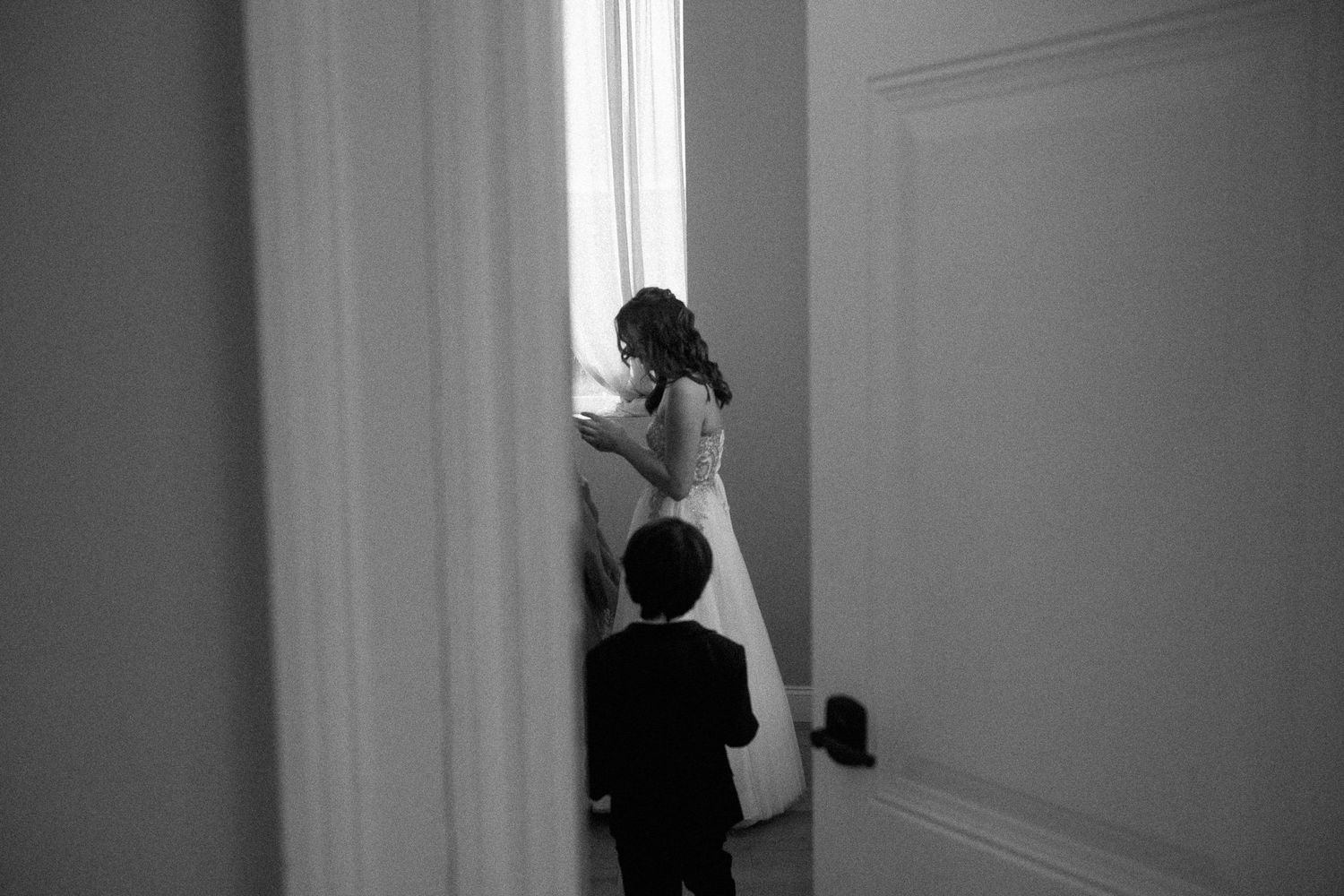 Black and white photo of a bride in a white gown standing by a window with a young boy in a suit beside her.