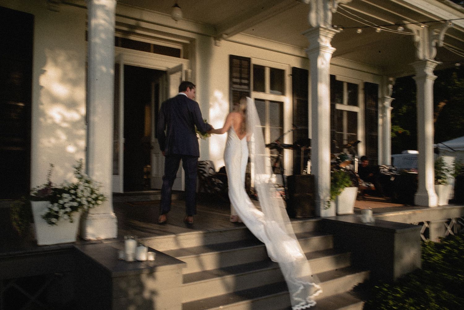 A bride and groom in wedding attire hold hands ascending porch steps of an elegant white colonial venue.