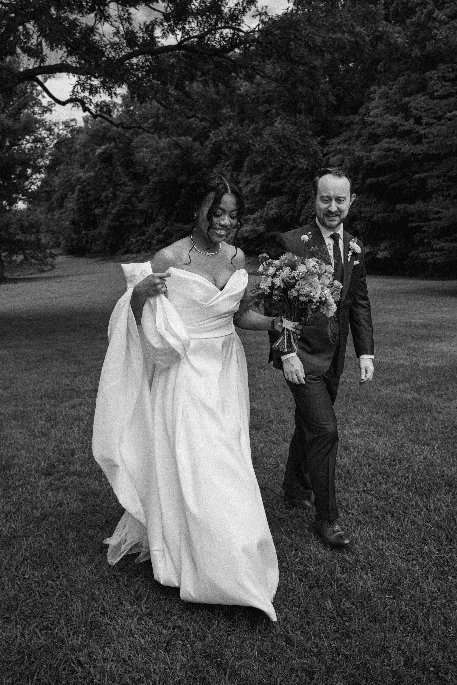 Black and white photo of smiling bride and groom walking across lush garden grounds on their wedding day.