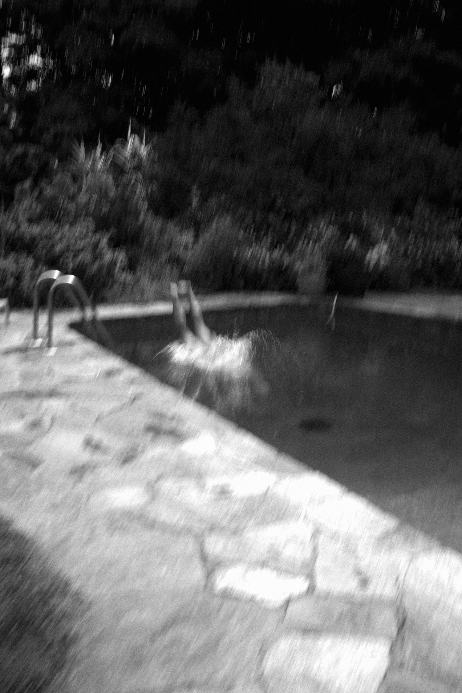 Blurry black and white photo of a stone-edged swimming pool with metal railings surrounded by lush greenery.