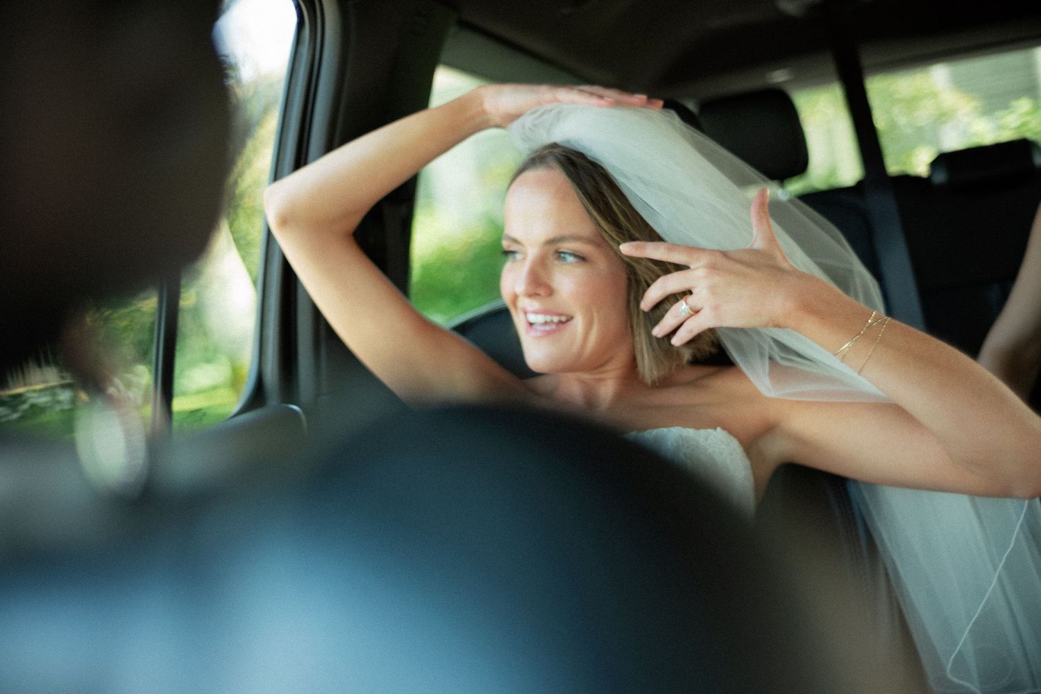 Smiling bride in veil adjusts hair while seated in car on wedding day.