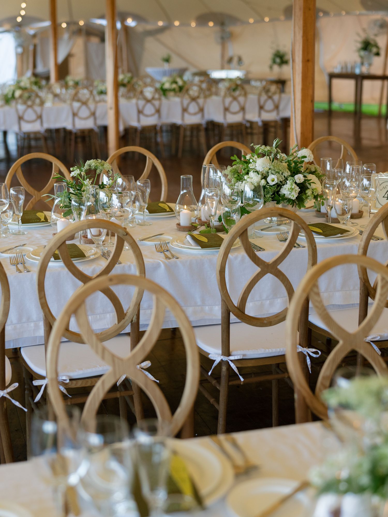 Wedding reception long tables with wooden infinity-back chairs, white floral centerpieces, and crystal glassware in a tent.