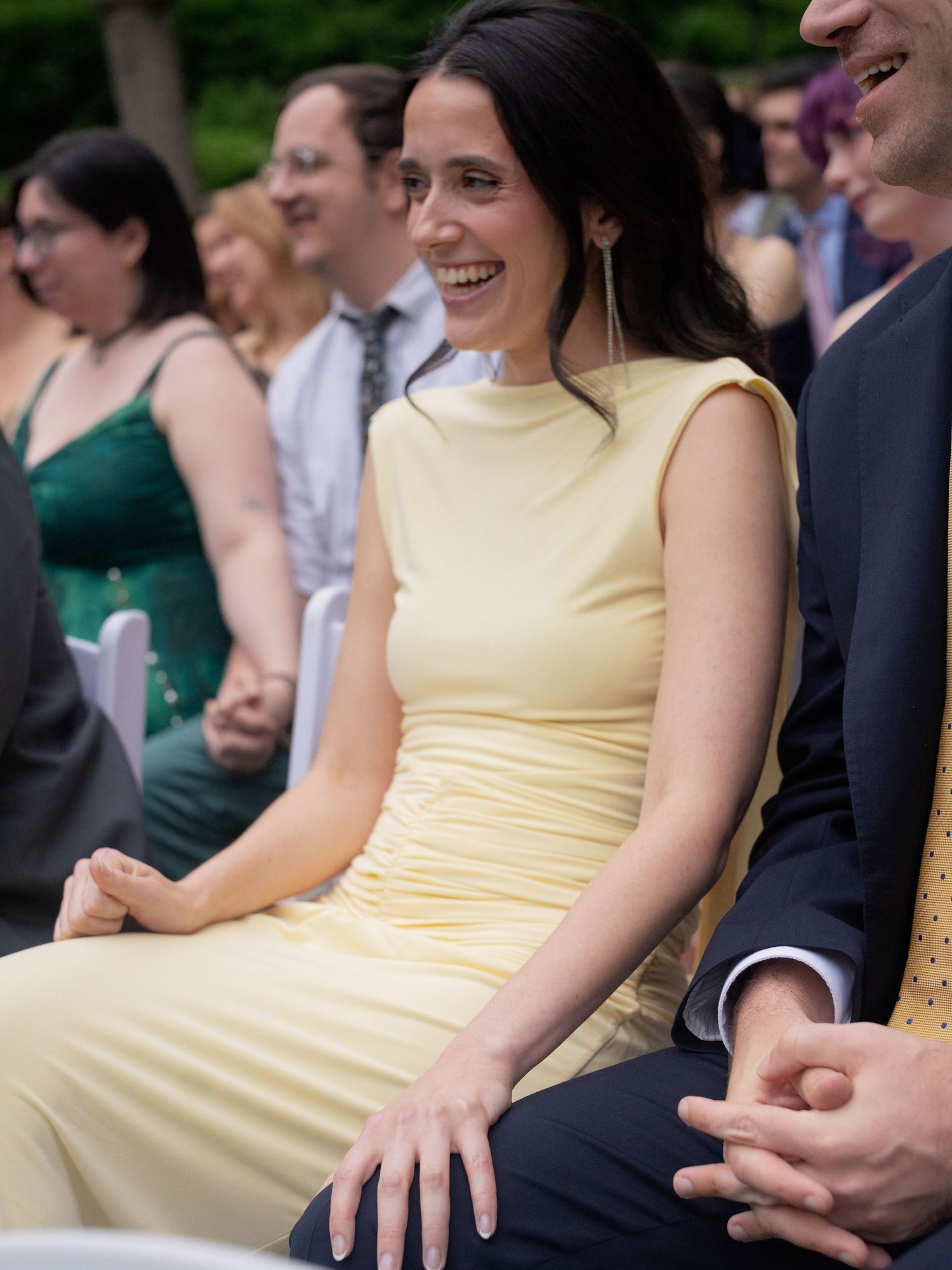 A smiling woman in a yellow dress enjoys an outdoor wedding ceremony as a guest.