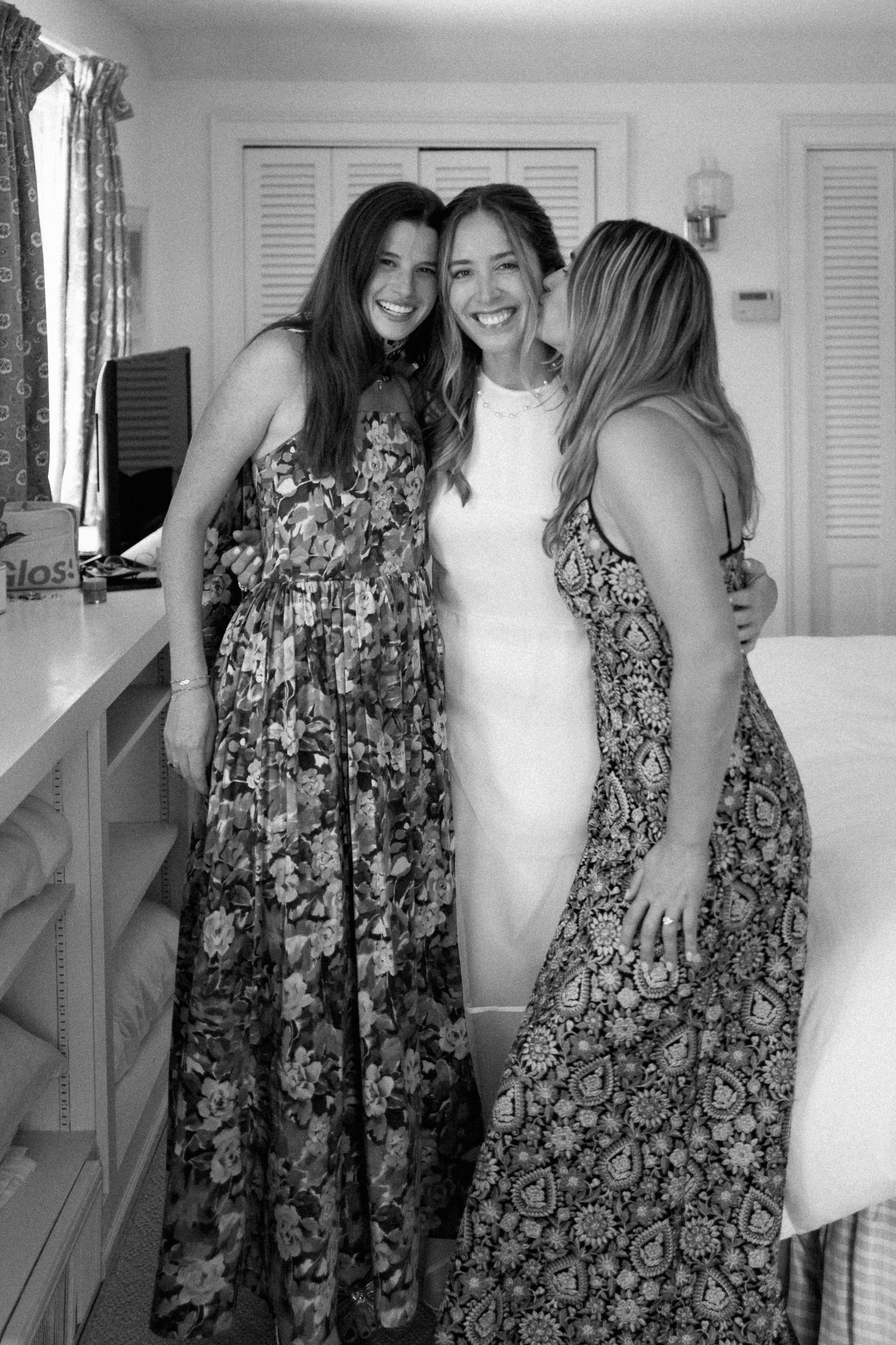 Bride in white gown shares joyful embrace with two friends in floral dresses on wedding day.