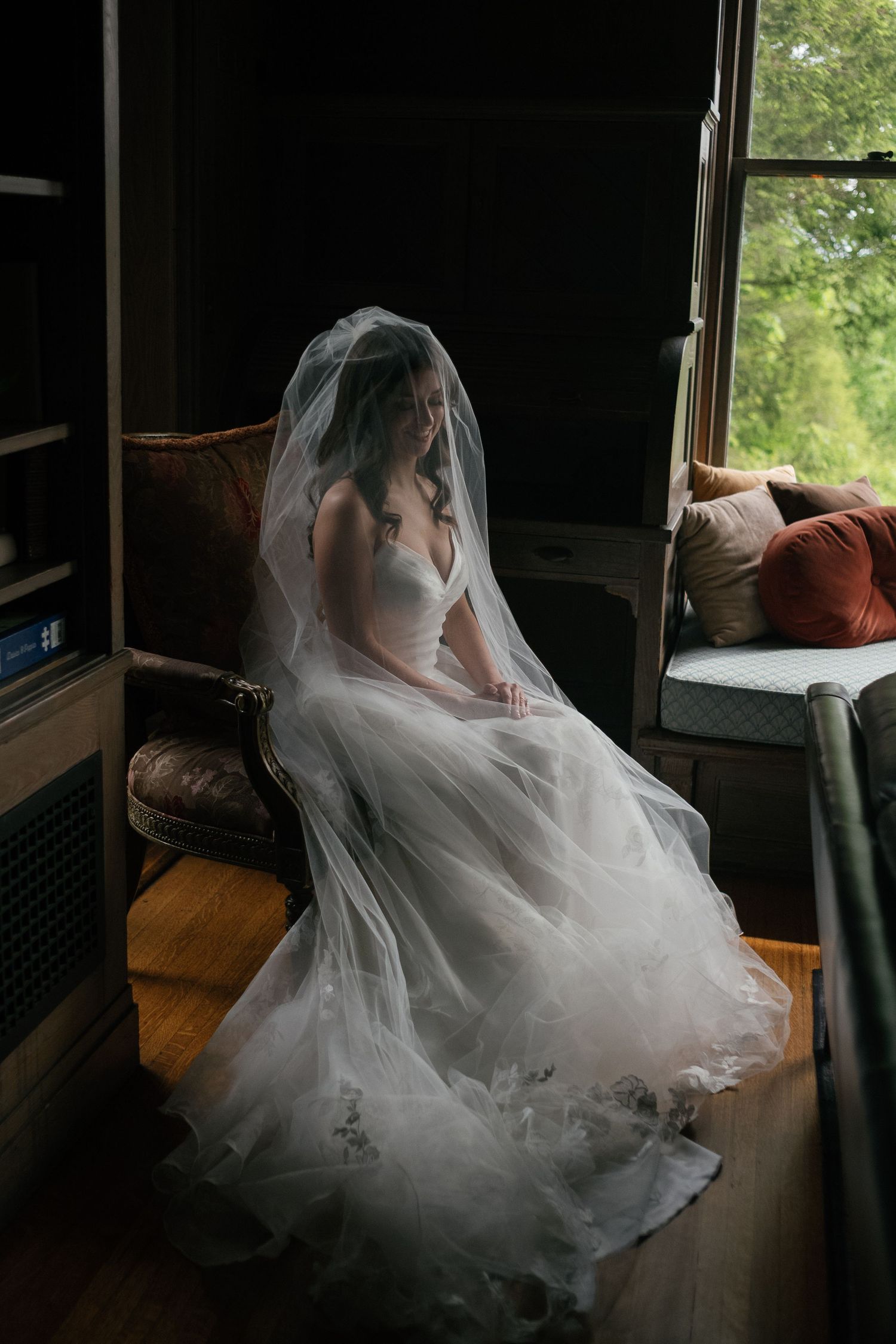Bride in a white gown and veil sits pensively by a window in a dimly lit vintage room.