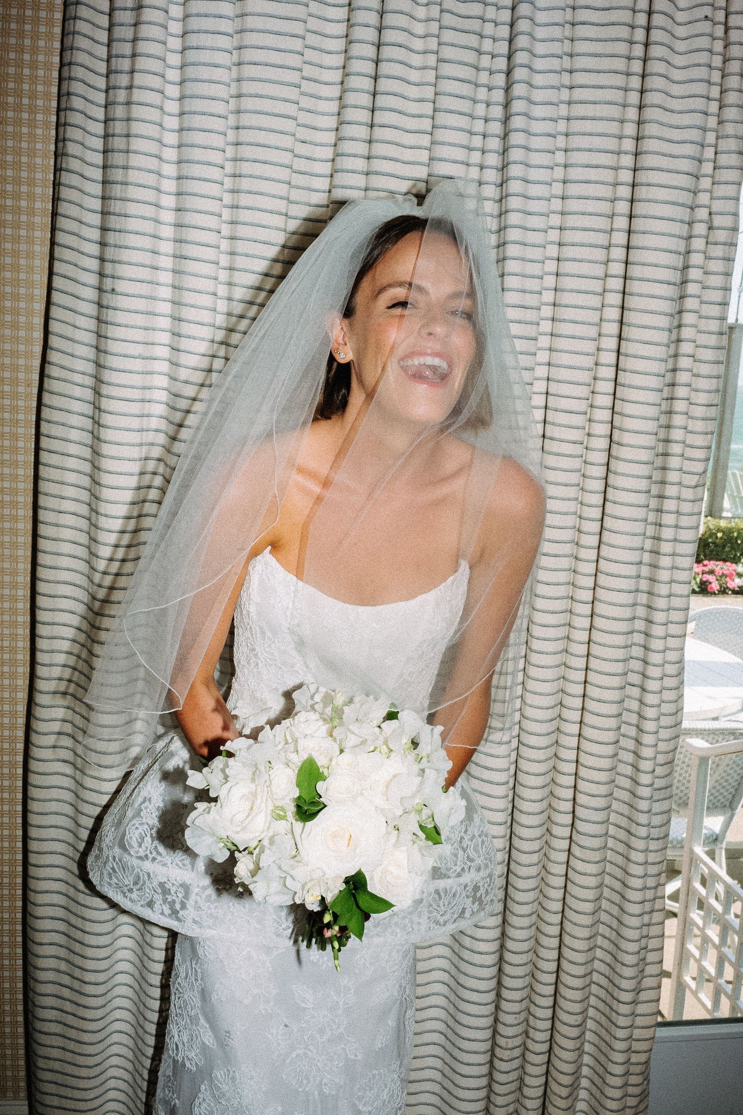 Smiling bride in white lace gown and veil holding white rose bouquet standing by striped curtains.