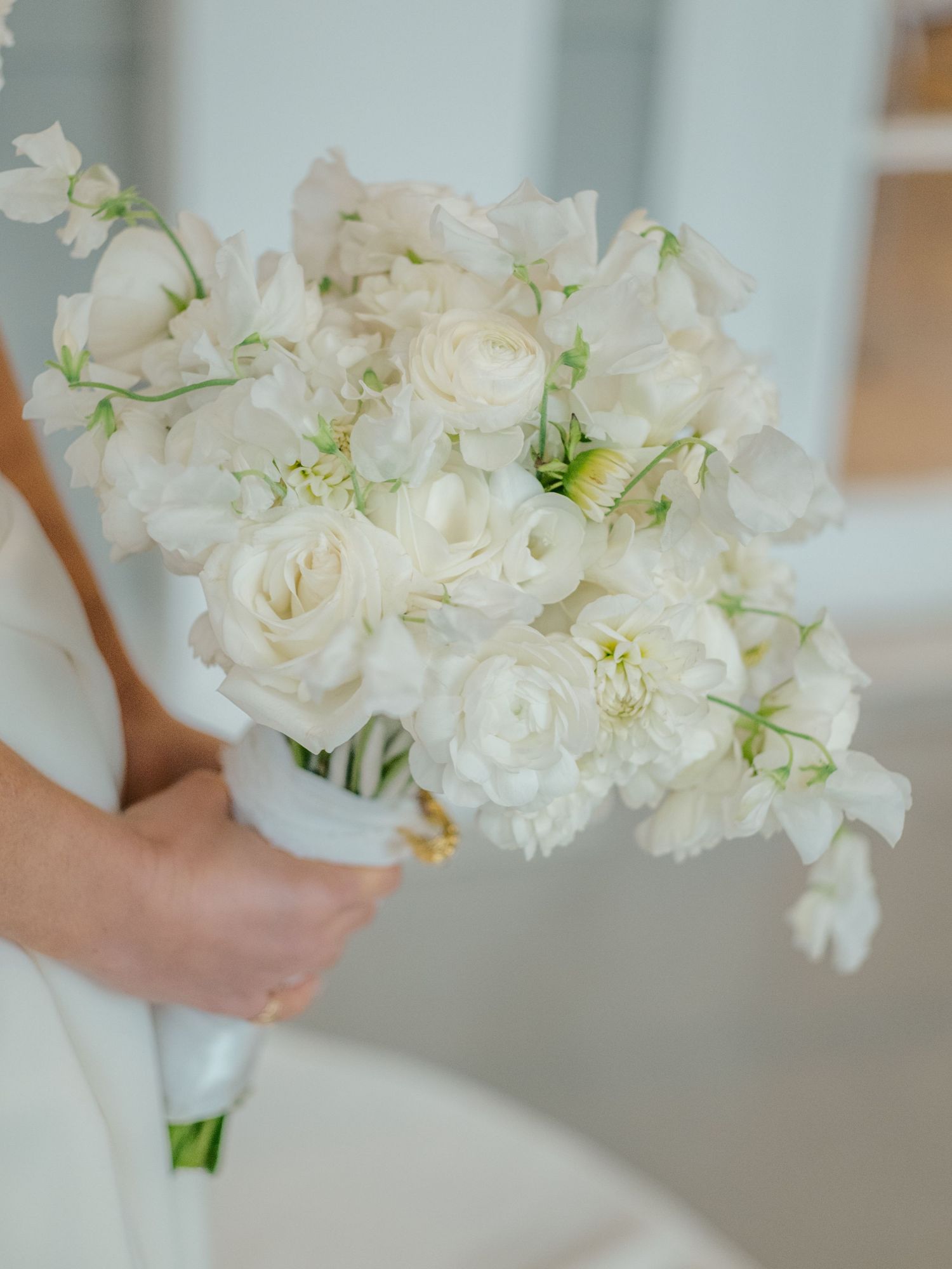 Elegant white wedding bouquet featuring ranunculus, roses, and sweet peas with lush greenery held by a bride.