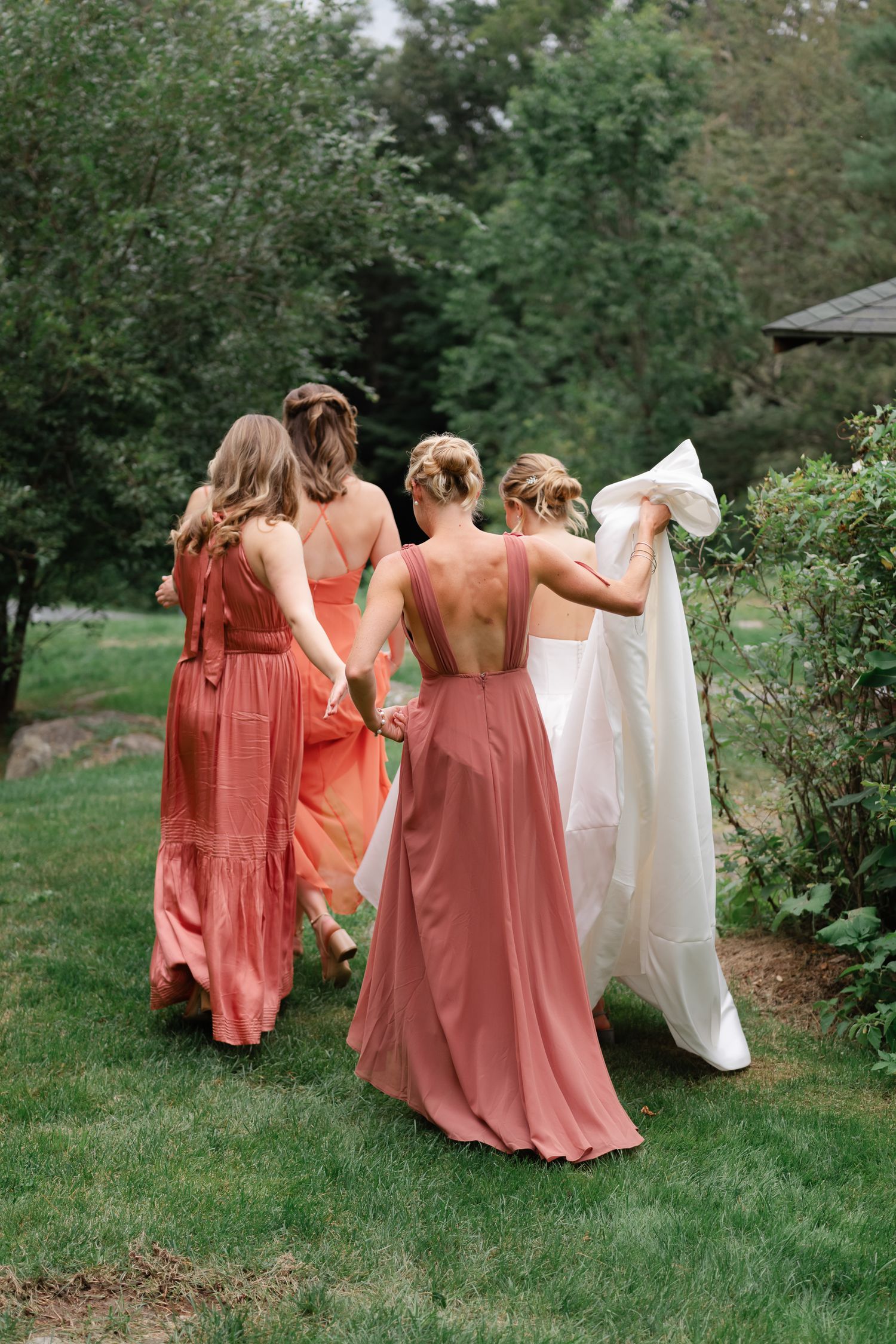 Bride and bridesmaids in coral dresses walk hand in hand through lush garden.