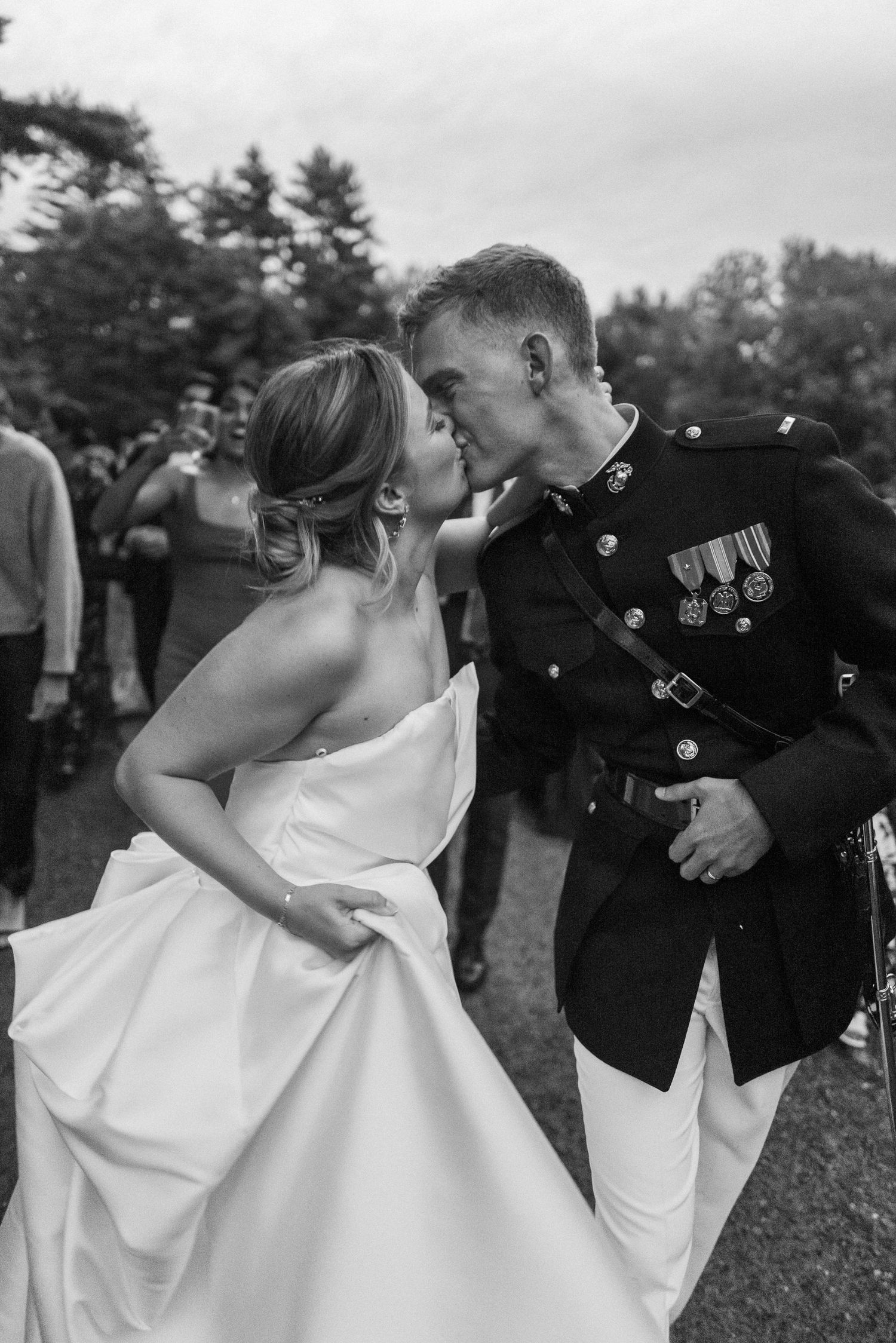 Newlywed couple kissing outdoors, groom in Marine dress blues with military medals, bride in elegant white gown.
