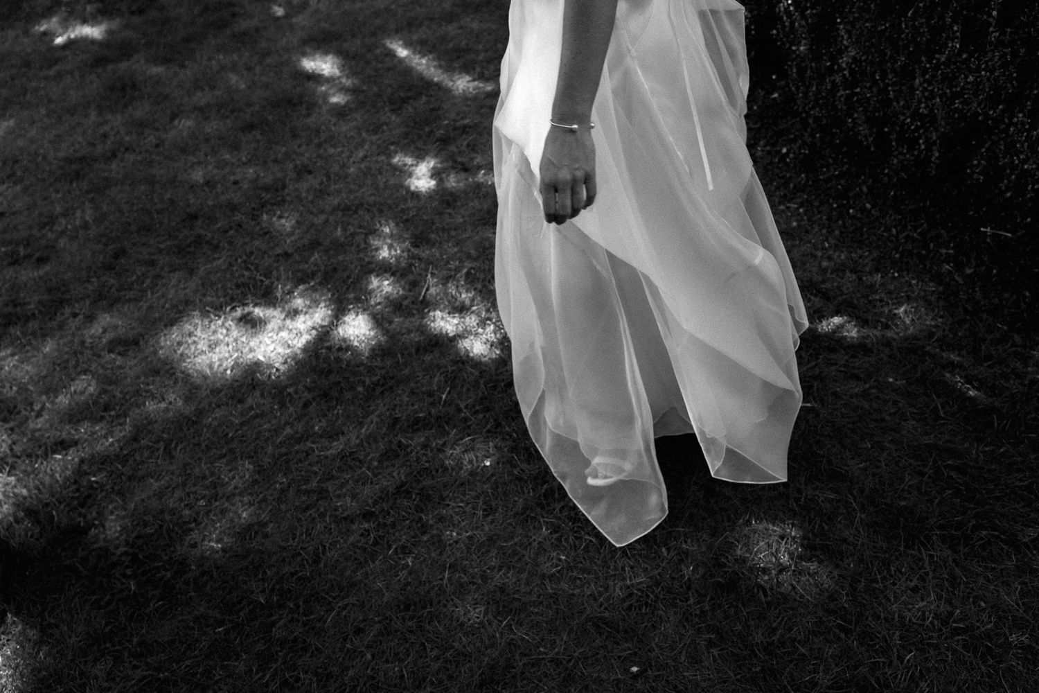 Black and white photo of bride in flowing white wedding gown standing on grass near a tree.