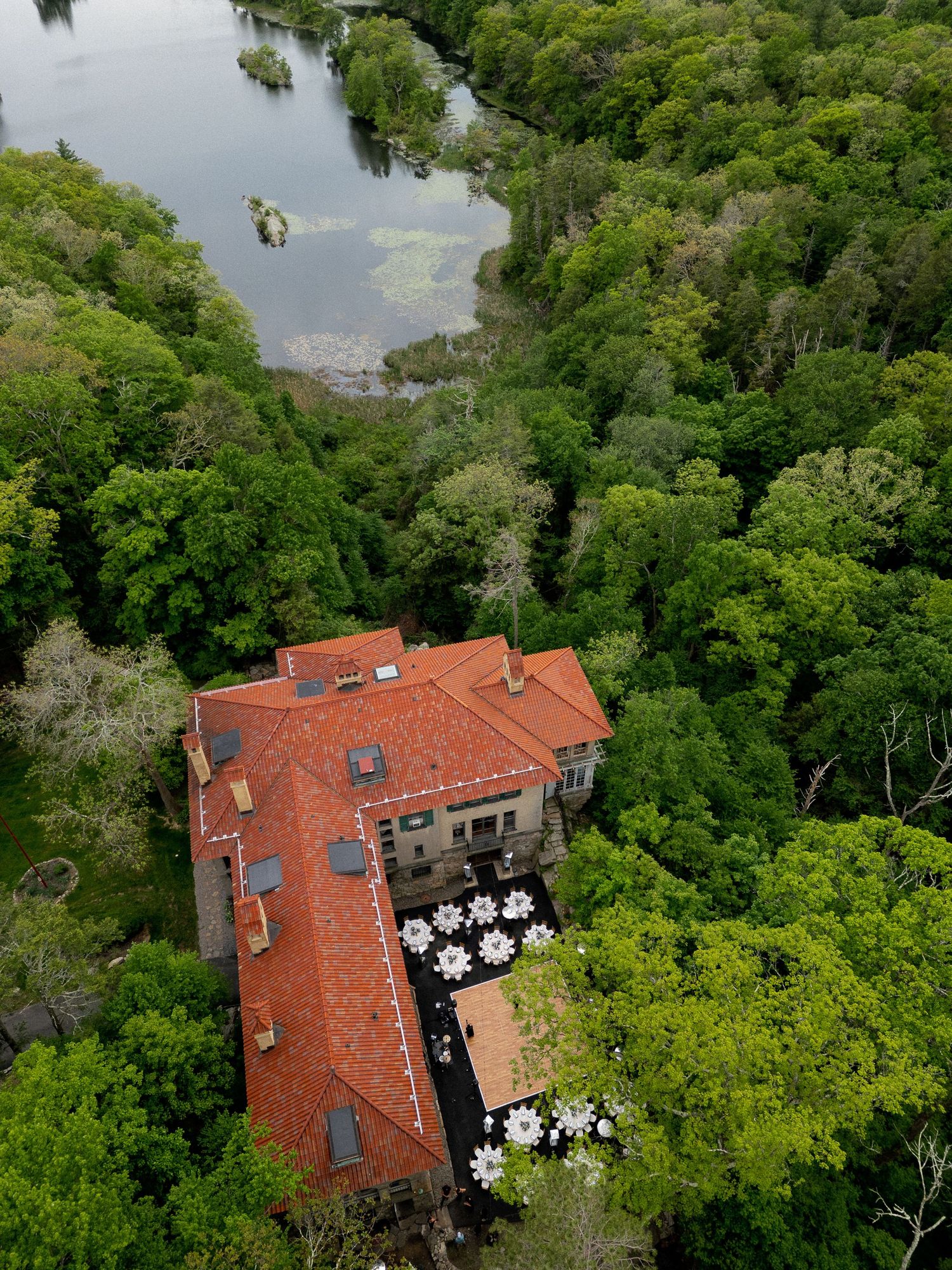 Aerial drone view of a stone mansion with red roof surrounded by lush trees near a lake.