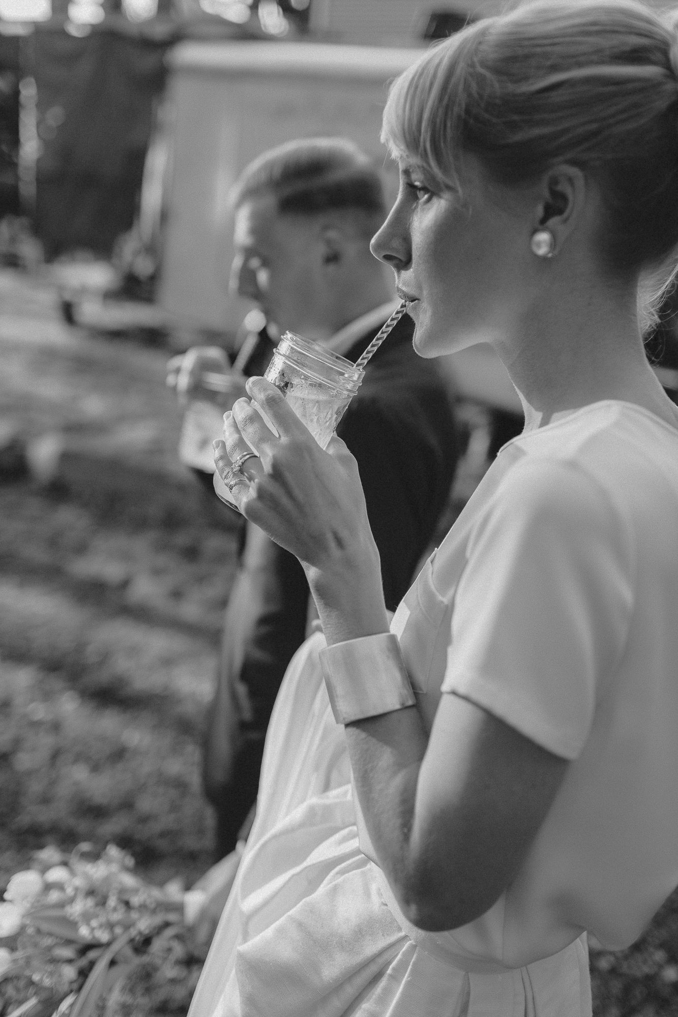 Black and white photo of a bride in white dress sipping a drink through a straw at an outdoor wedding reception.