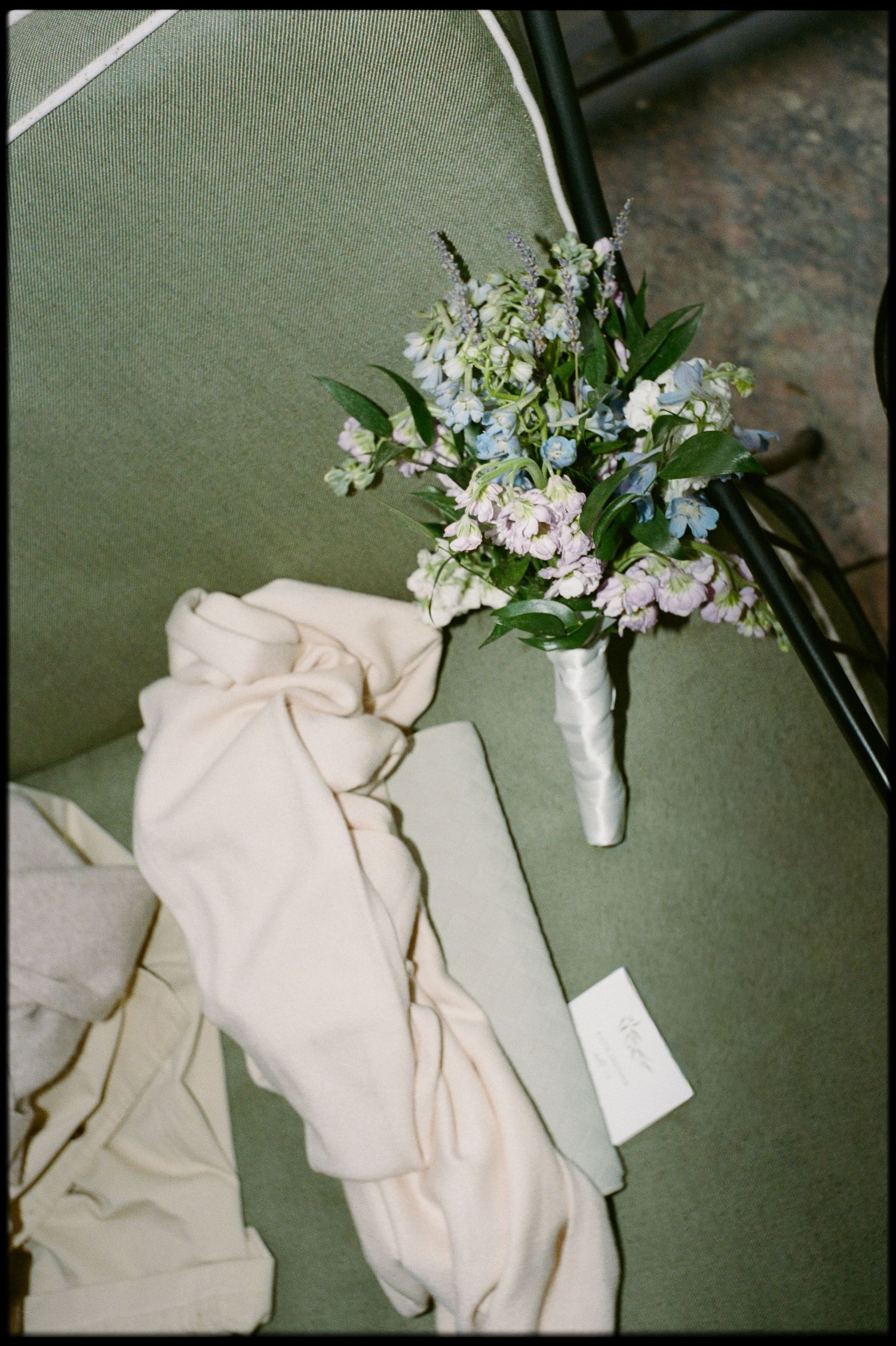 Bridal bouquet with blue, purple, and white wildflowers resting on green chair beside flowing white fabric.
