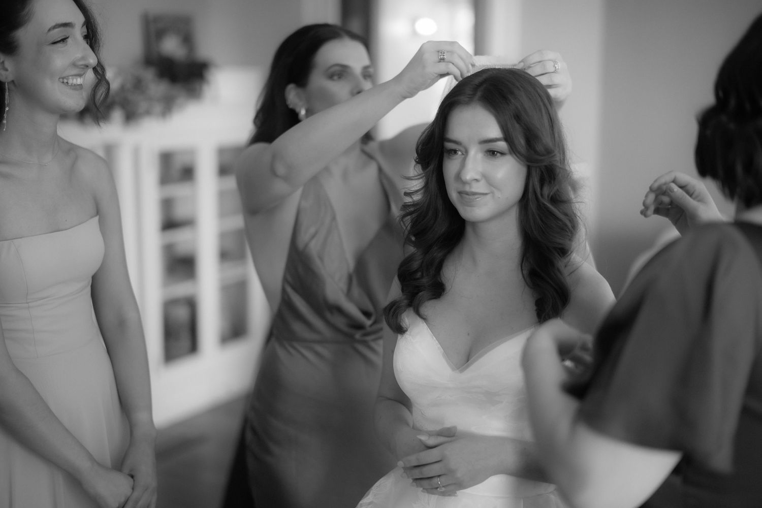 Bridesmaids help bride with hair and dress during wedding preparations in bright room.
