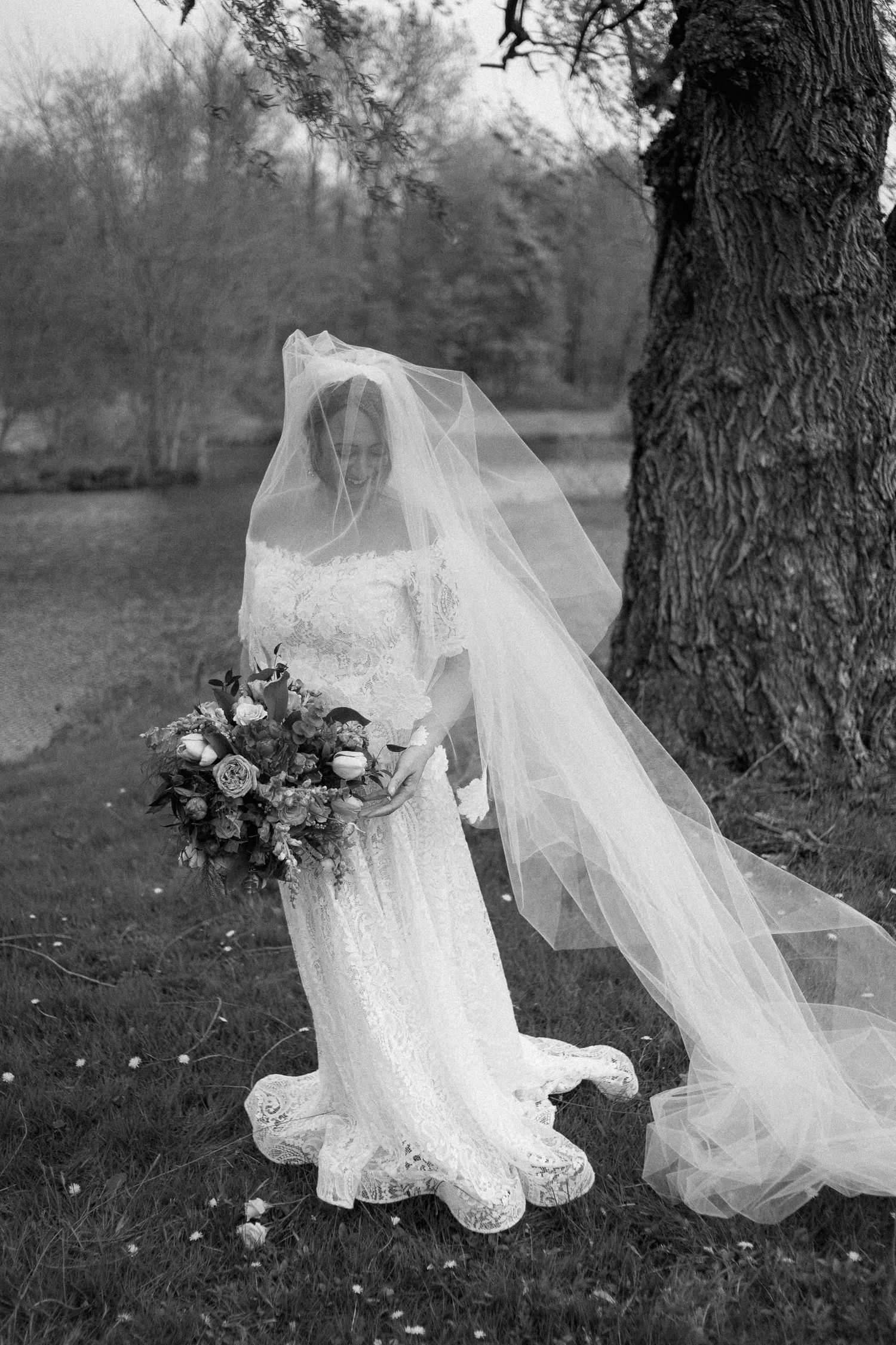 Black and white photo of a bride in lace gown and flowing veil holding bouquet outdoors near a large tree.
