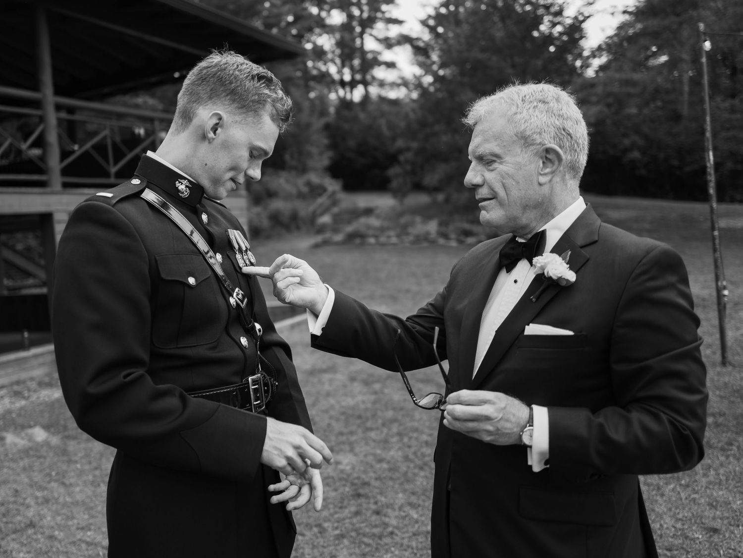 Marine groom in dress uniform shares moment with older man in tuxedo outdoors.