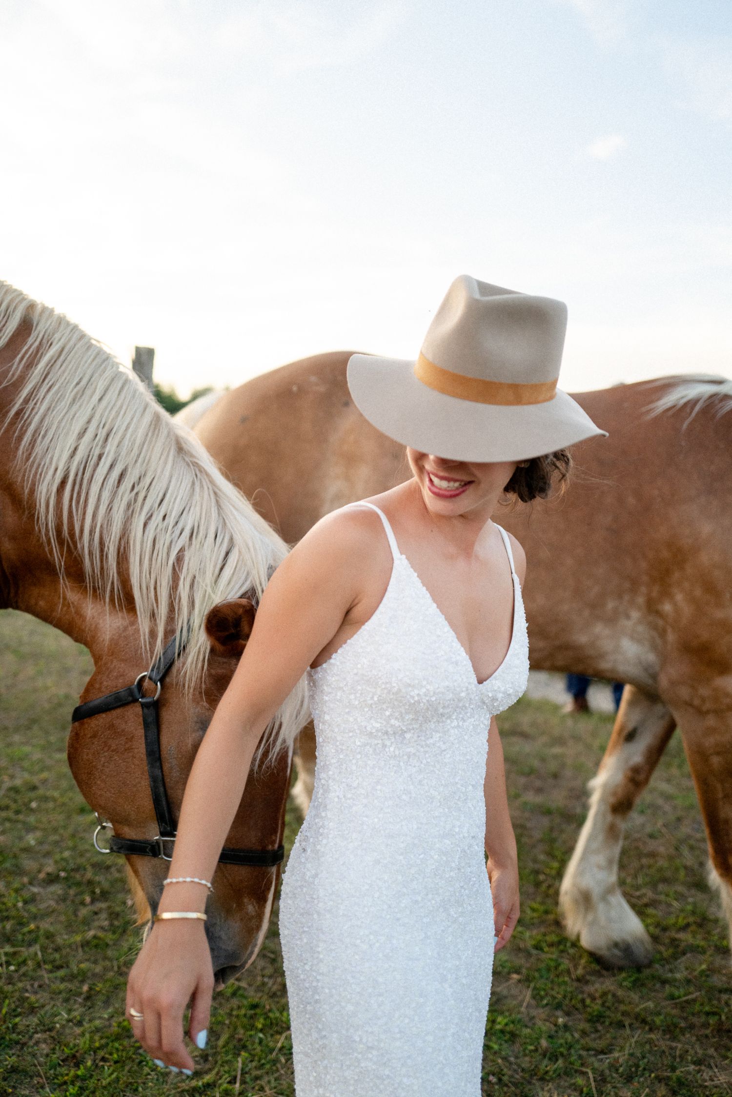 Laughing bride in white sequin gown and wide-brim hat poses with palomino horses at golden hour.