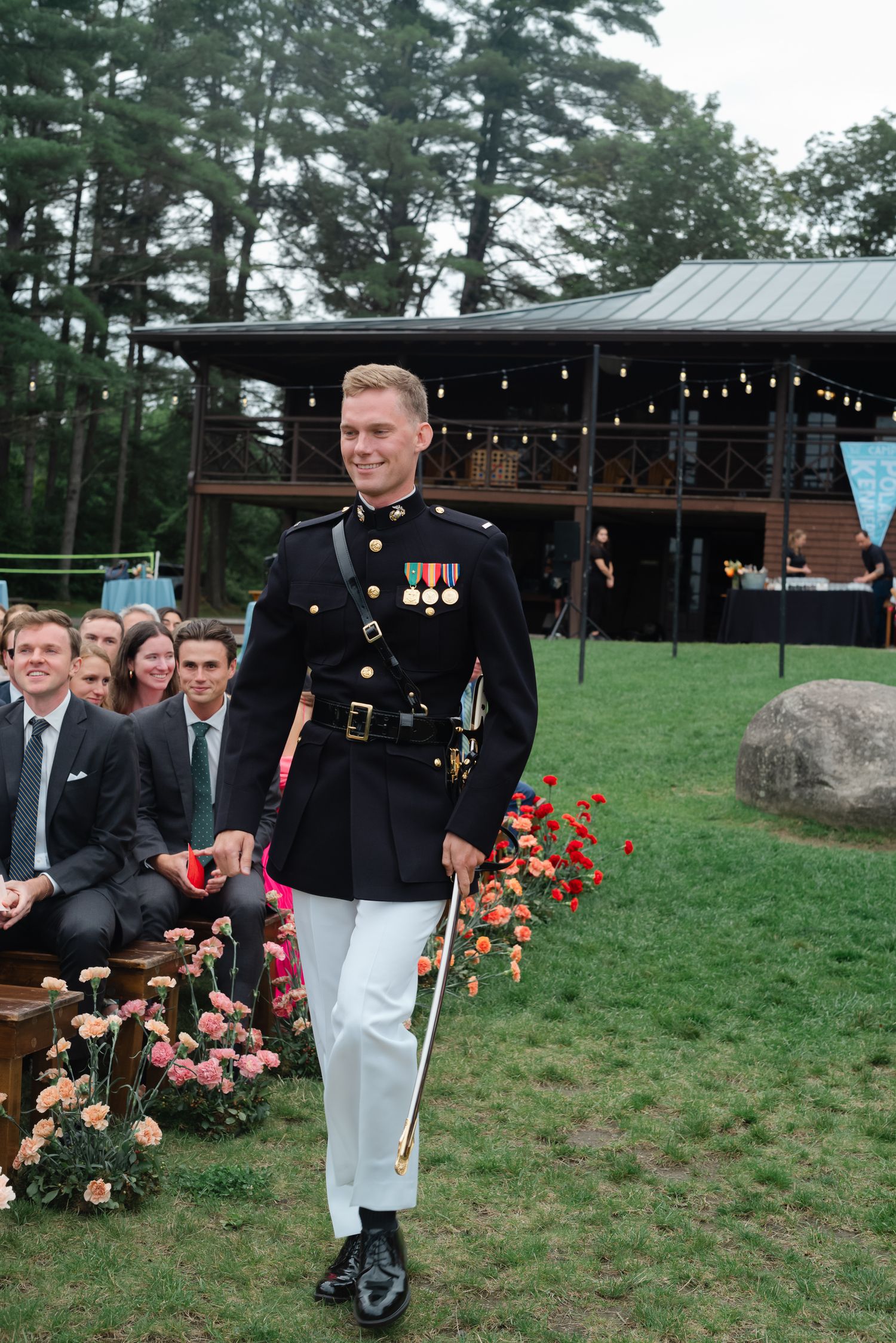 Groom in Marine dress uniform with medals walking down outdoor wedding ceremony aisle with floral arrangements.