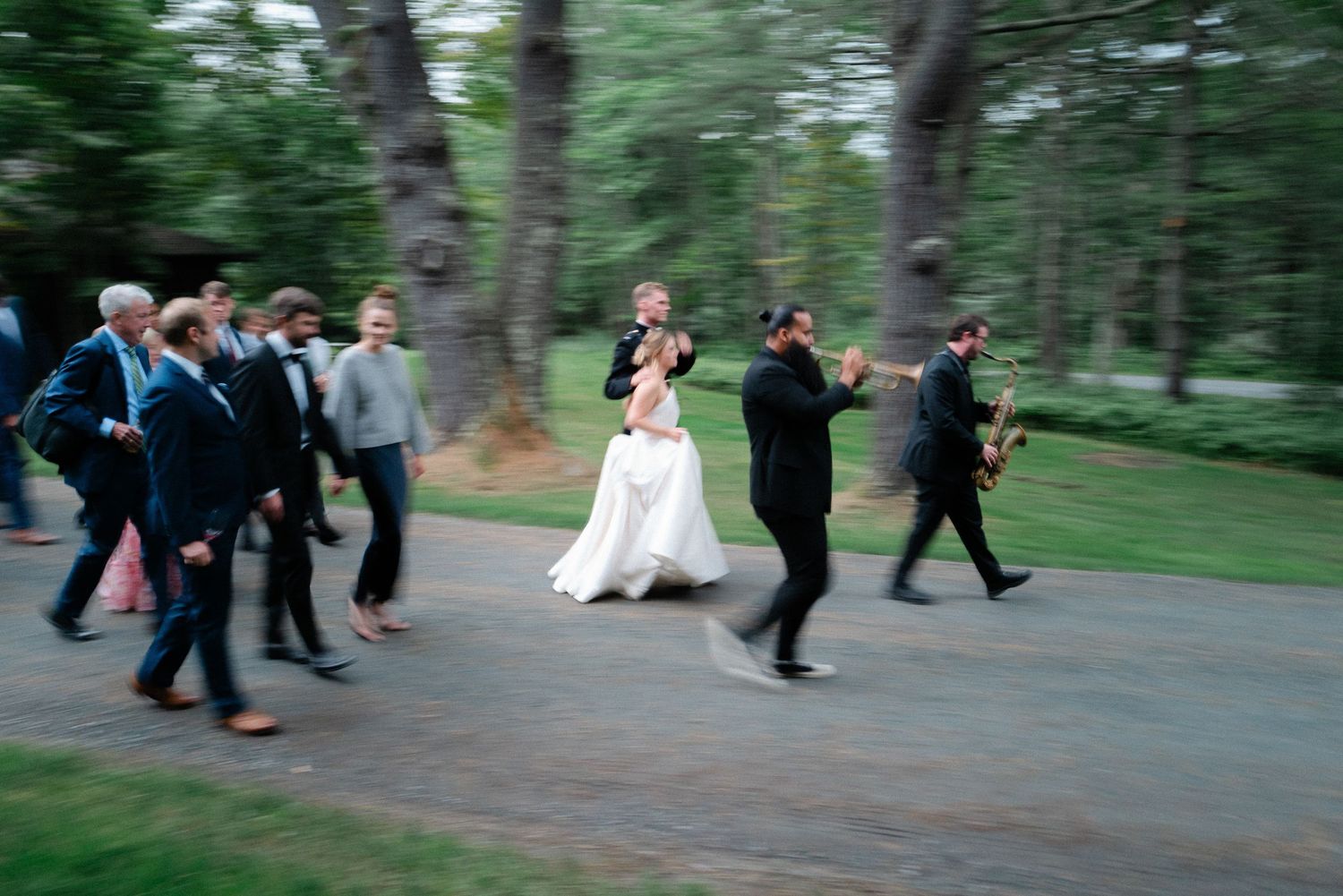 Motion-blurred wedding party processional through pine tree lined path with bride in white gown and guests.