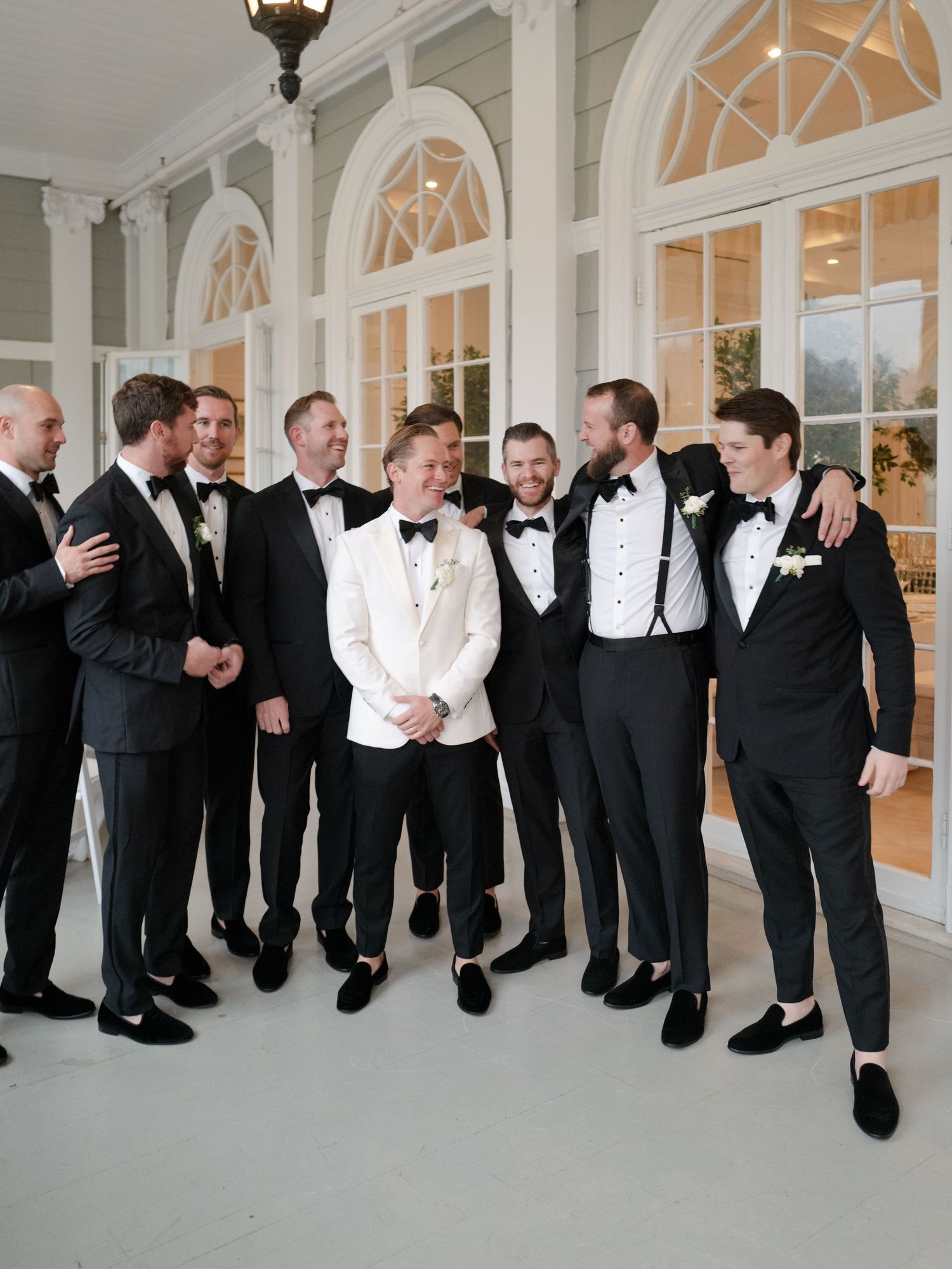 Groom in white tuxedo jacket laughing with groomsmen in black tuxedos outside elegant venue.
