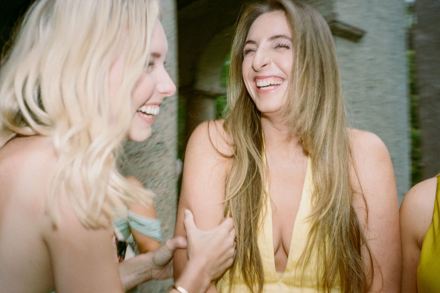 Two women laughing and smiling together outdoors, one in a yellow dress, sharing a joyful moment.