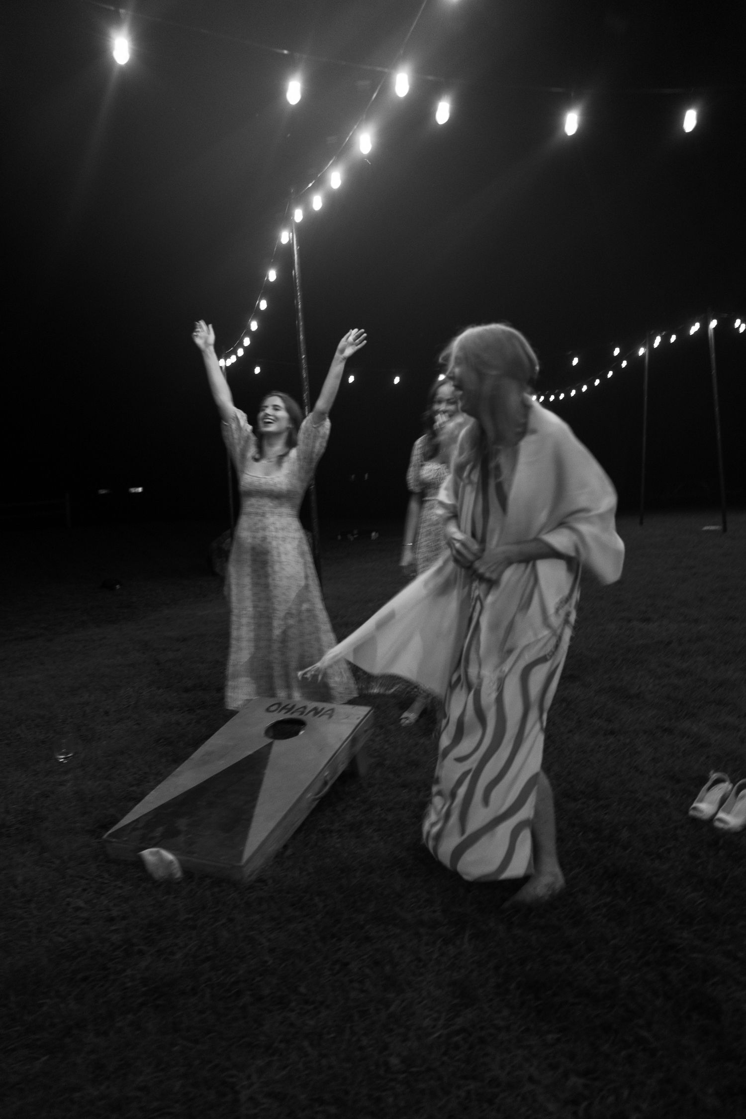 Black and white photo of women playing cornhole under string lights at a nighttime outdoor wedding.