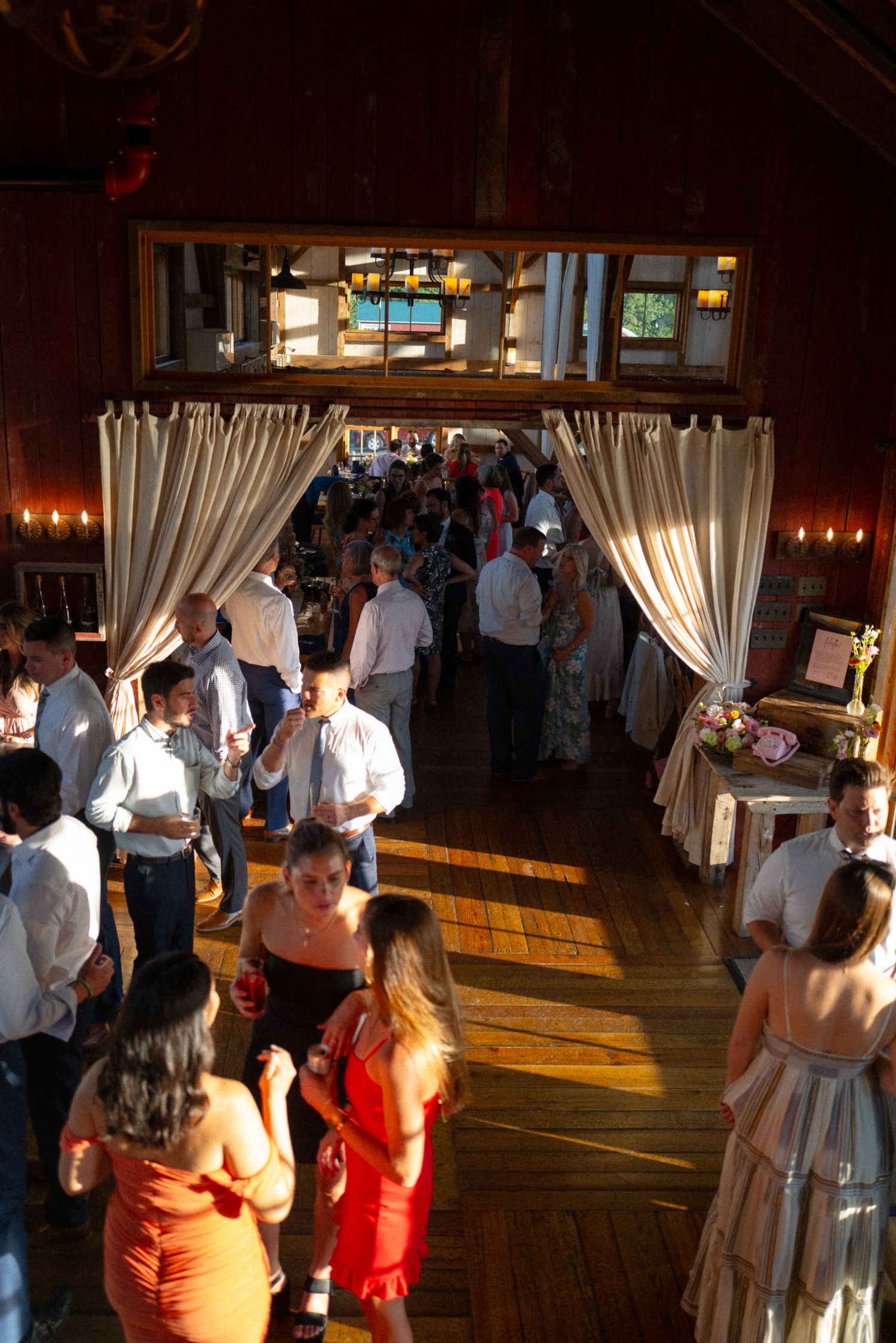 Wedding guests mingle and dance in a warmly lit rustic barn reception hall with elegant draping.