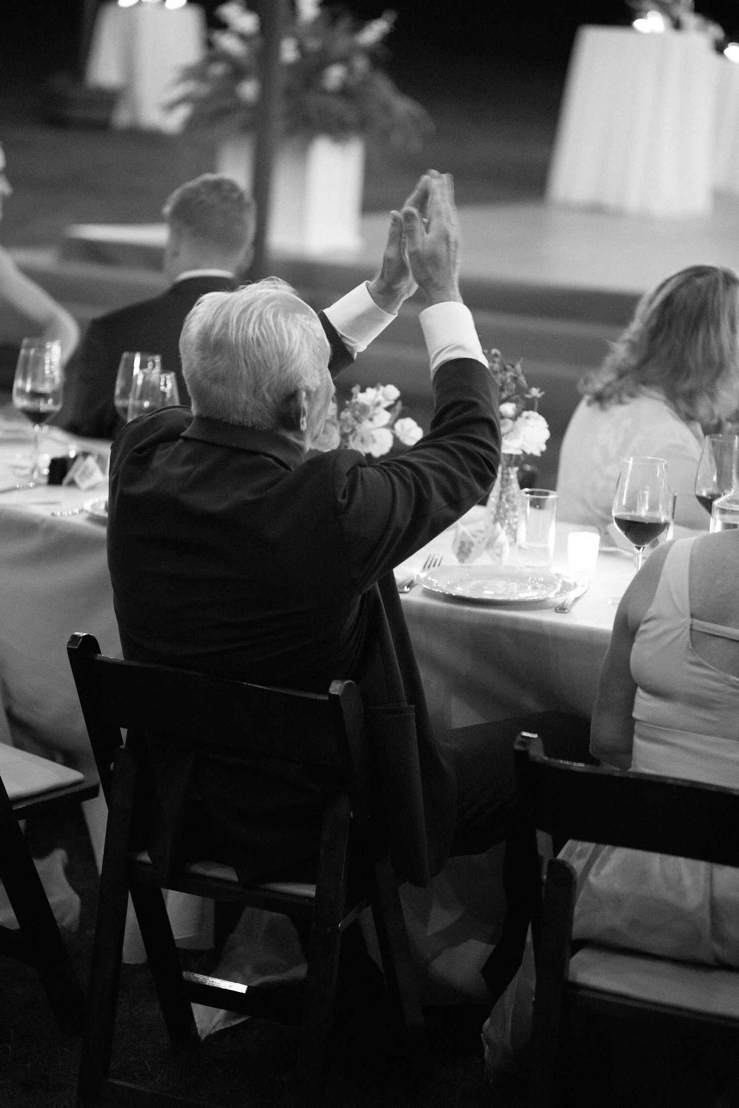 Black and white photo of elderly wedding guest applauding at an outdoor reception dinner table with wine glasses.