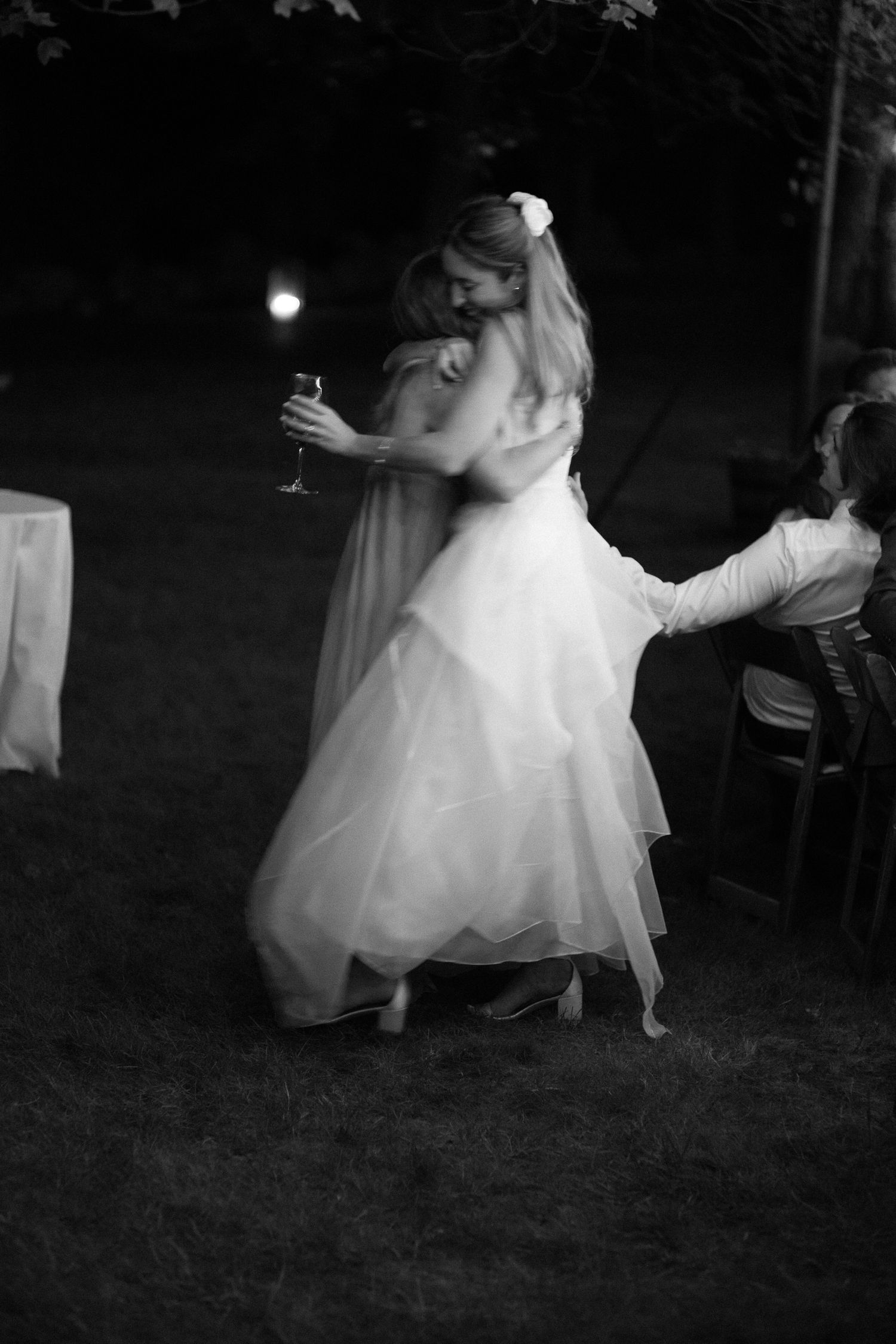 Black and white photo of bride in layered ball gown dancing outdoors at night holding a champagne glass.