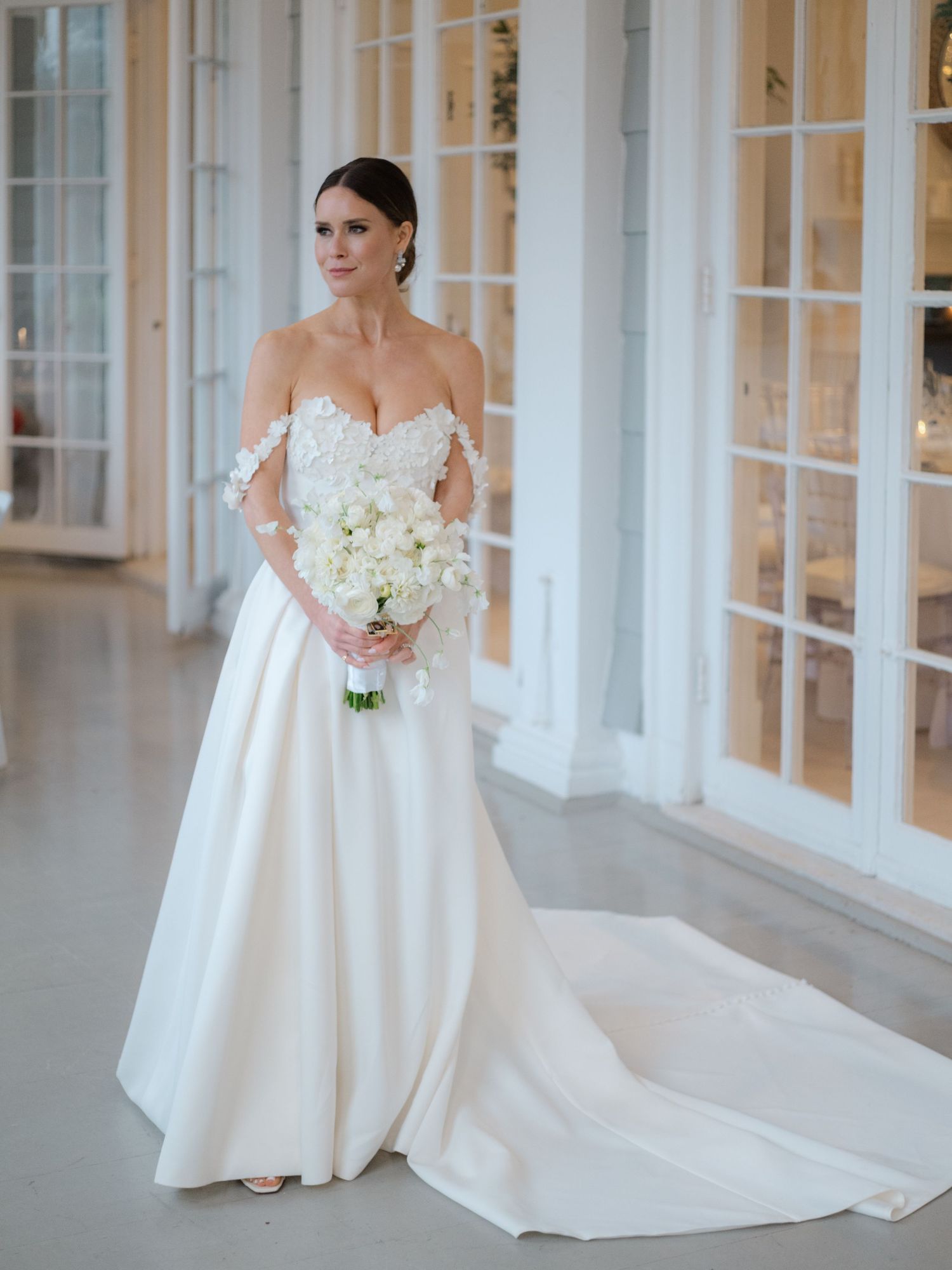 Elegant bride in off-shoulder lace wedding gown holds white orchid bouquet in sunlit venue.