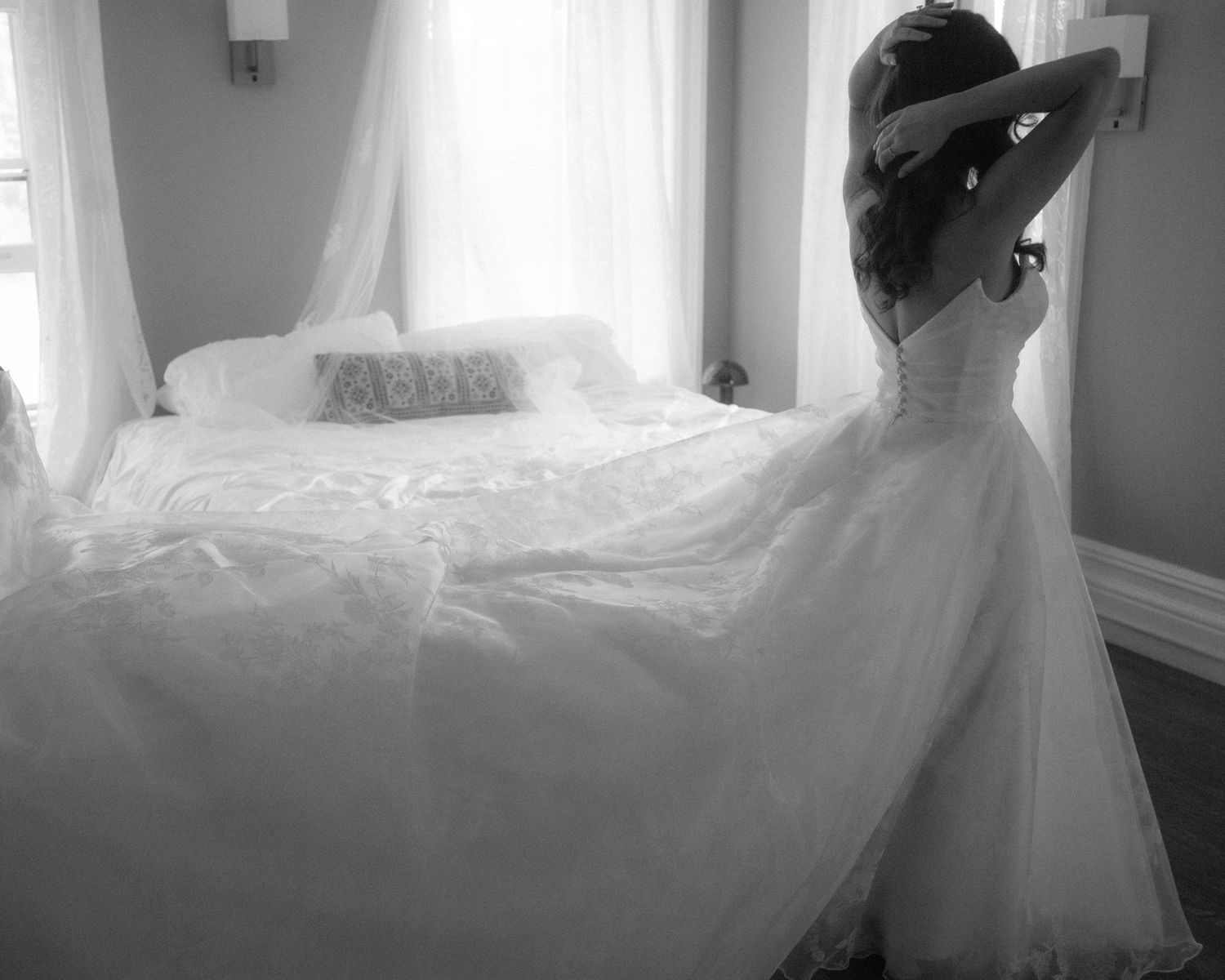 Black and white artistic photo of a bride in a full white wedding gown standing near a sunlit window.