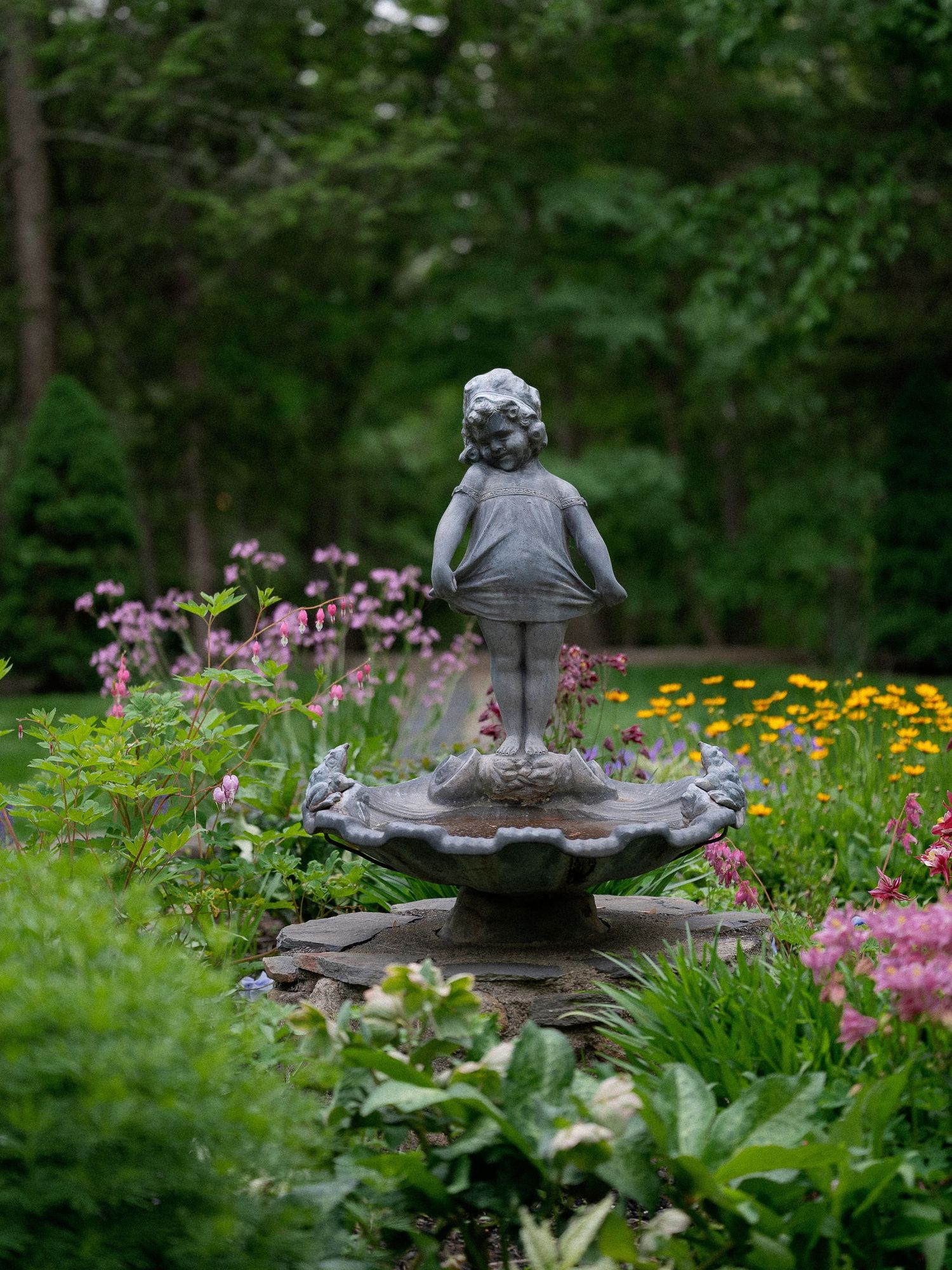 Stone fountain statue of a child surrounded by colorful blooming flowers in a garden.