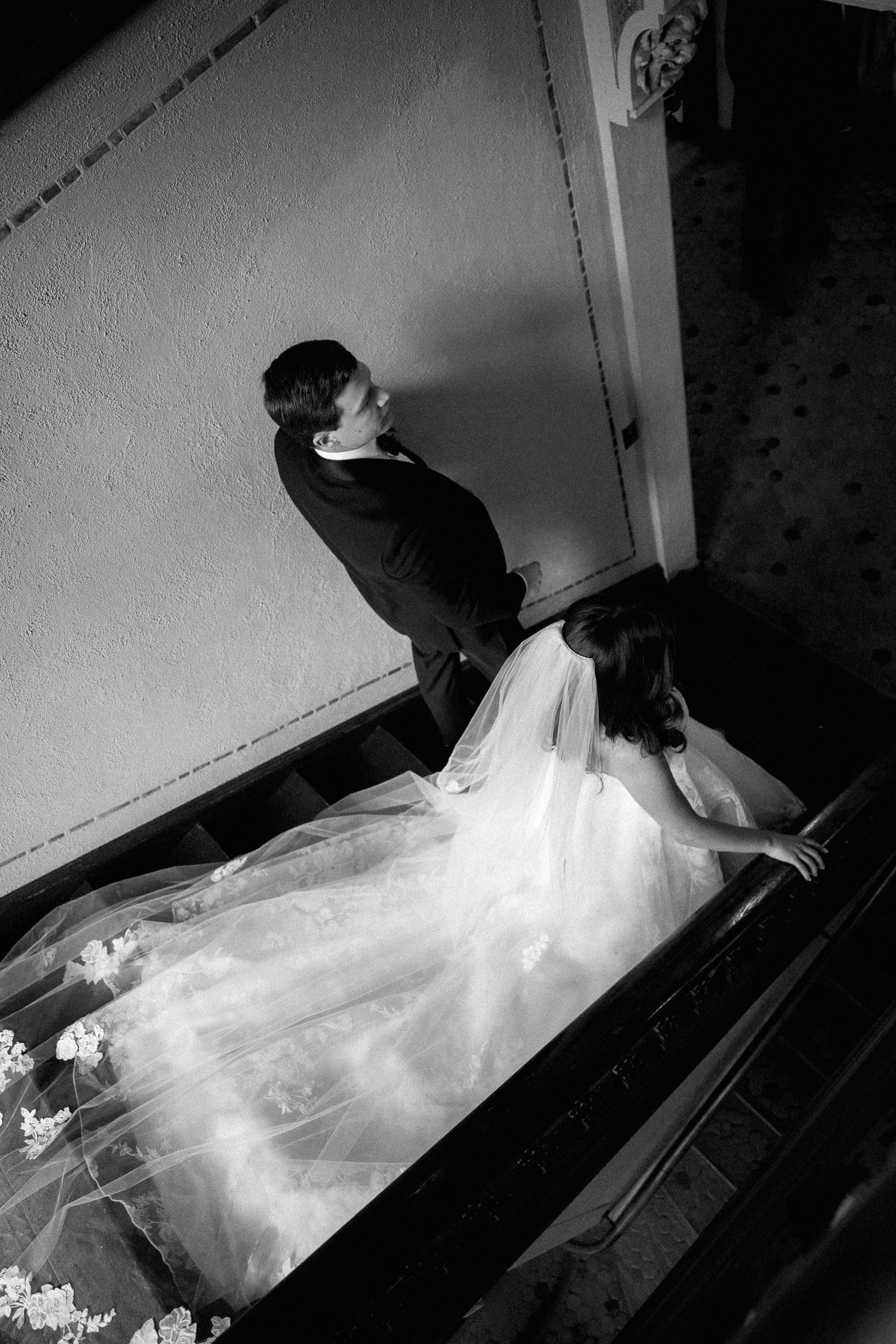 Black and white overhead photo of bride in flowing gown and groom on grand staircase at wedding venue.