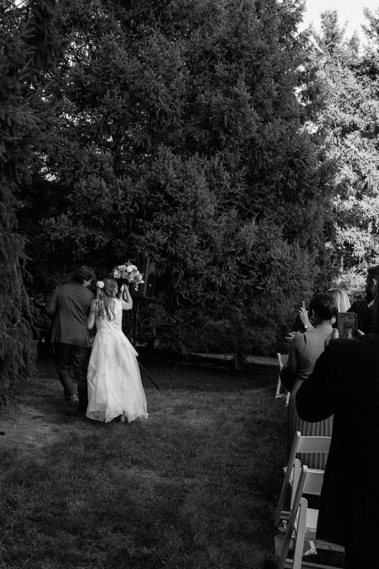 Black and white photo of bride being escorted down the outdoor aisle toward the wedding ceremony.