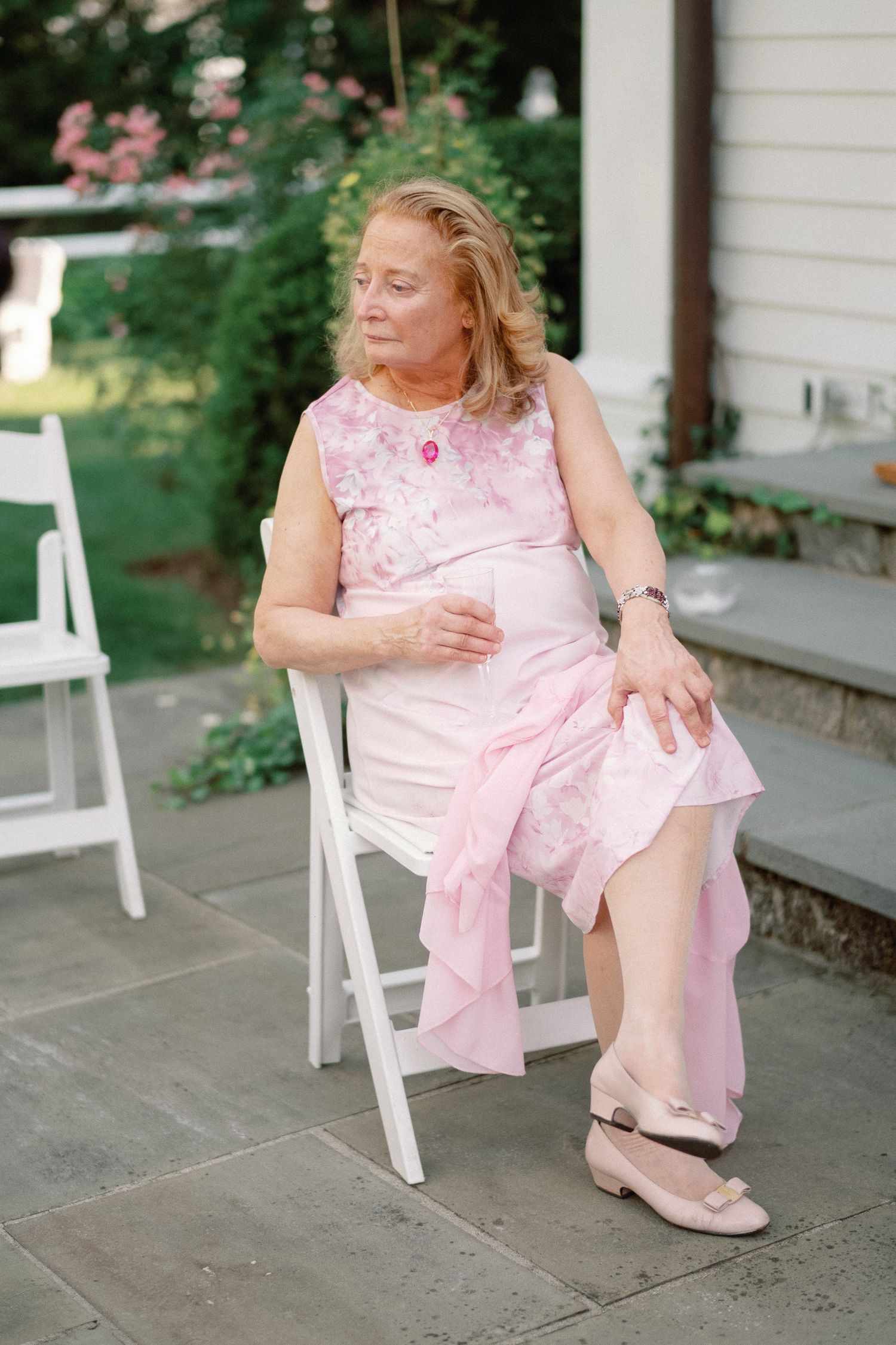 Elegant older woman in pink floral dress seated on white chair at outdoor wedding.