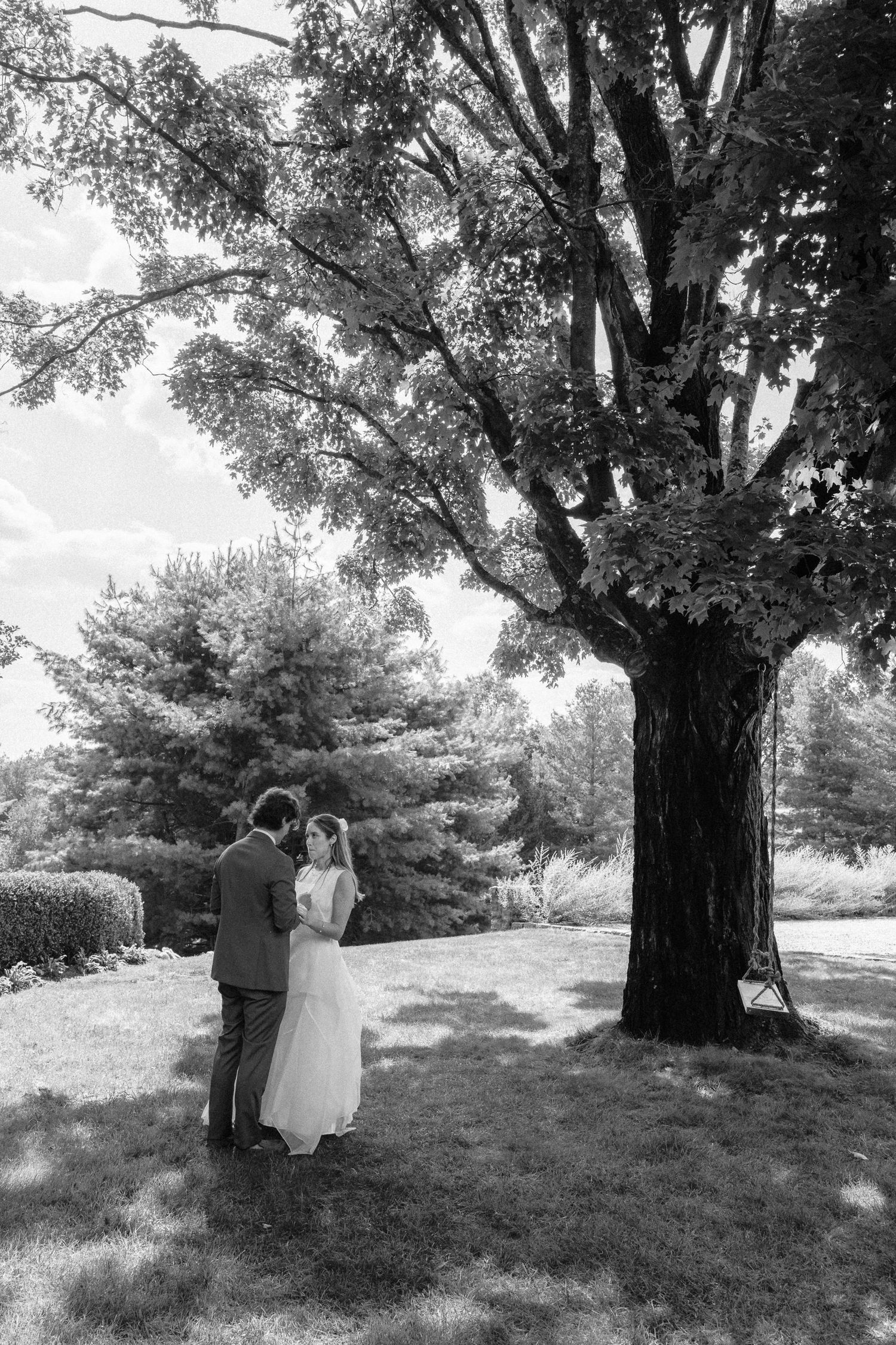 Black and white photo of bride and groom embracing under a large tree in a lush garden setting.