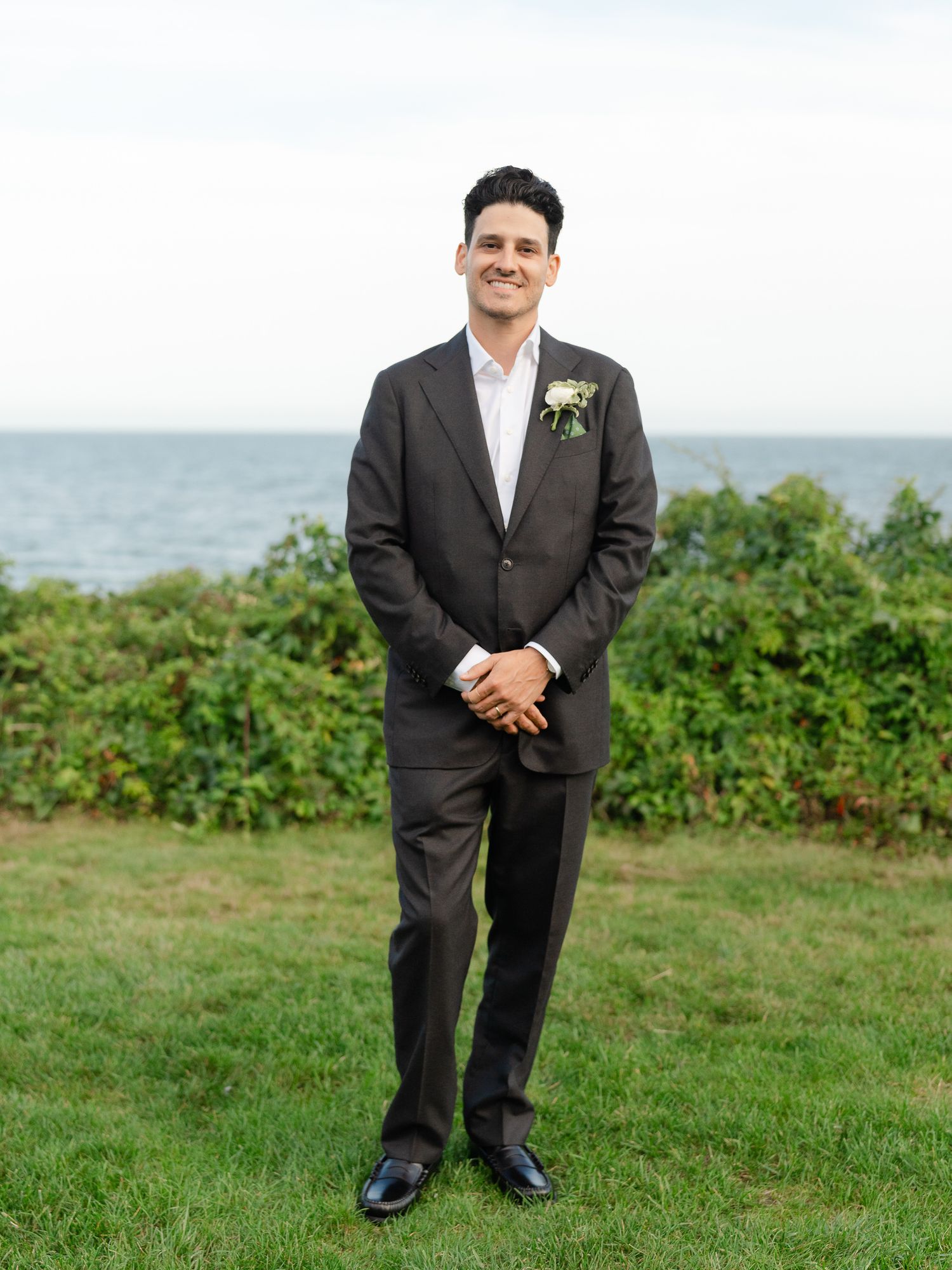 Handsome groom in dark suit with boutonniere stands before ocean backdrop at wedding.