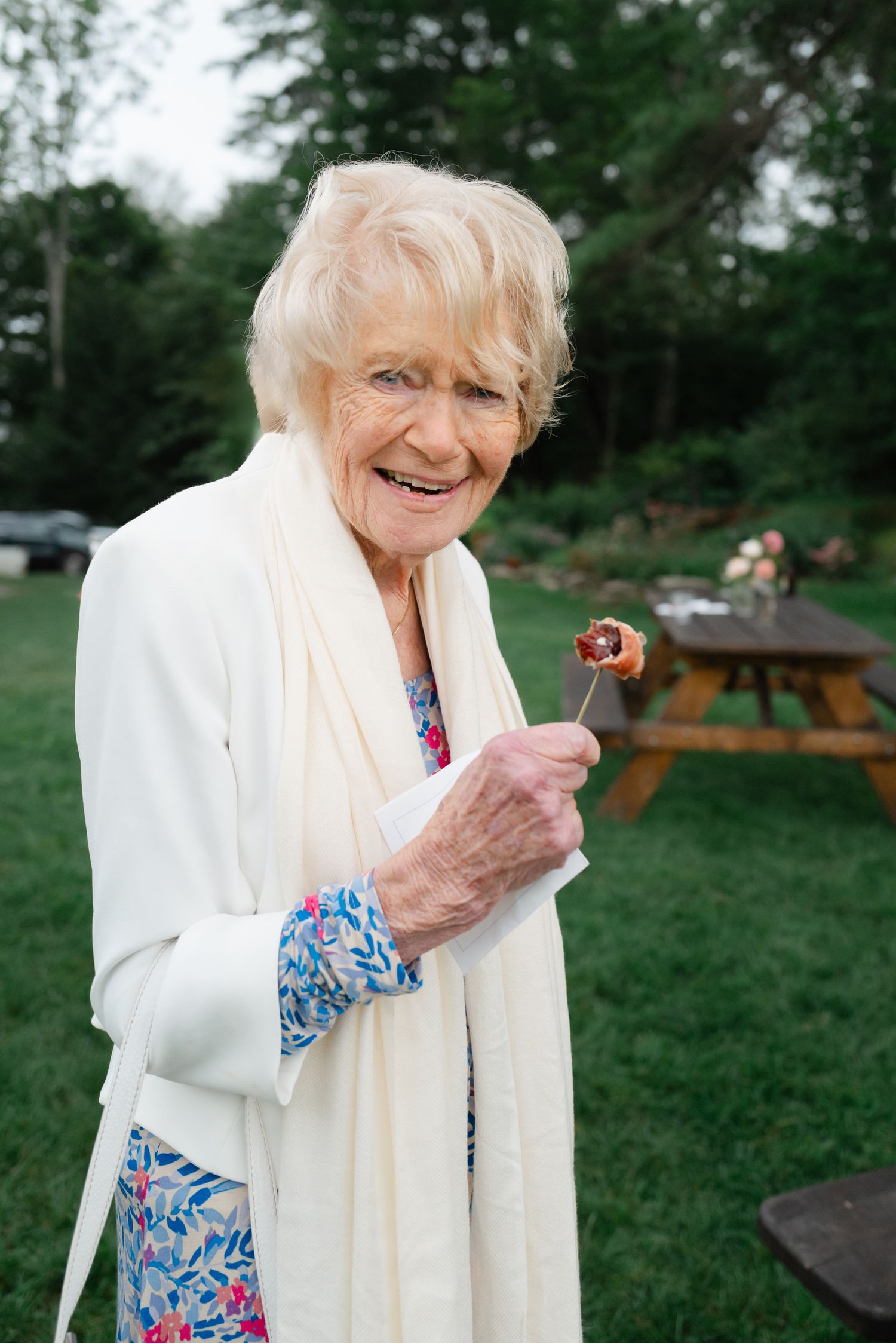Smiling elderly woman in white shawl and floral dress enjoying food at an outdoor wedding reception.