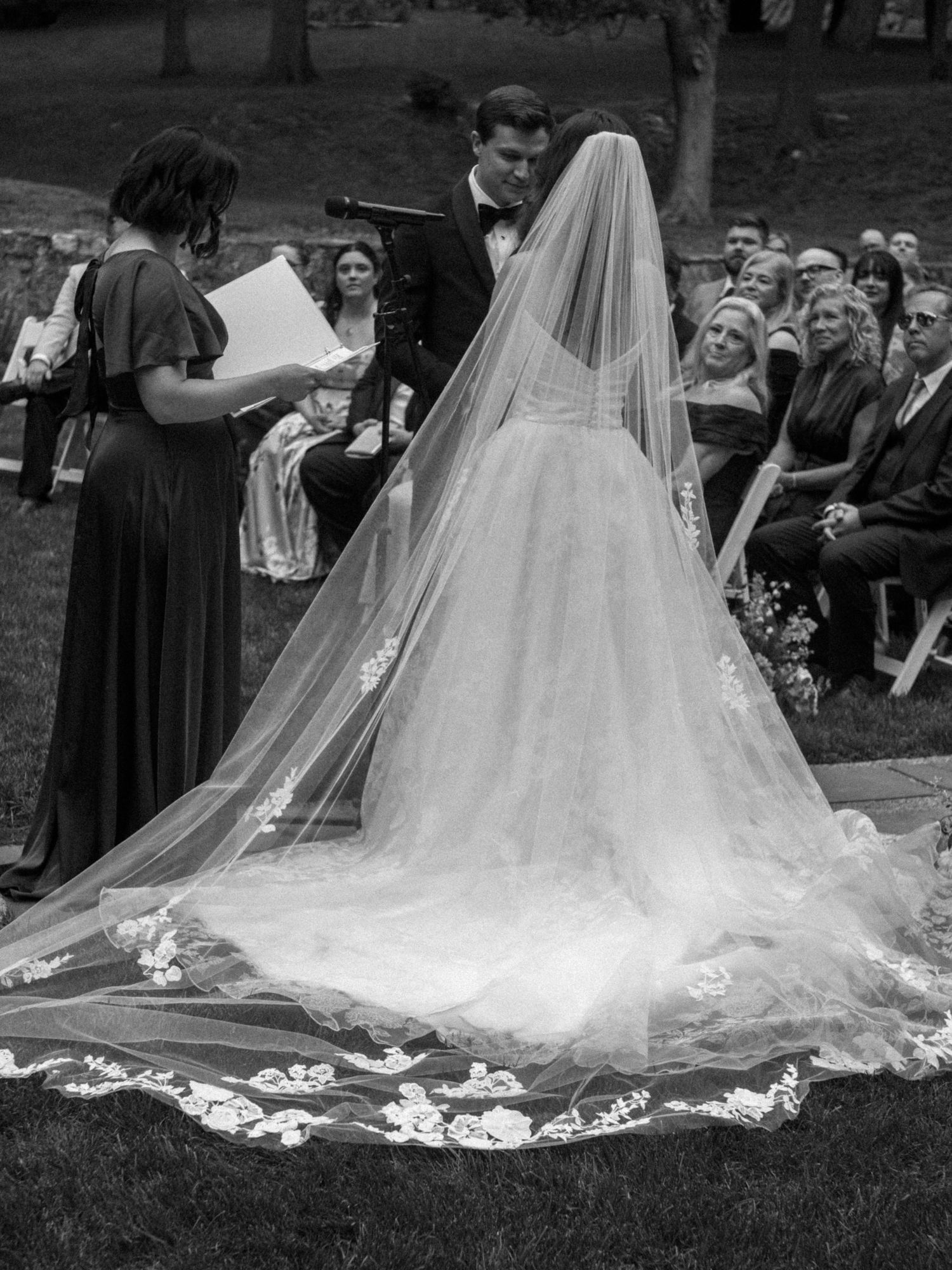Black and white photo of a bride in a lace-trimmed cathedral veil during outdoor wedding ceremony.