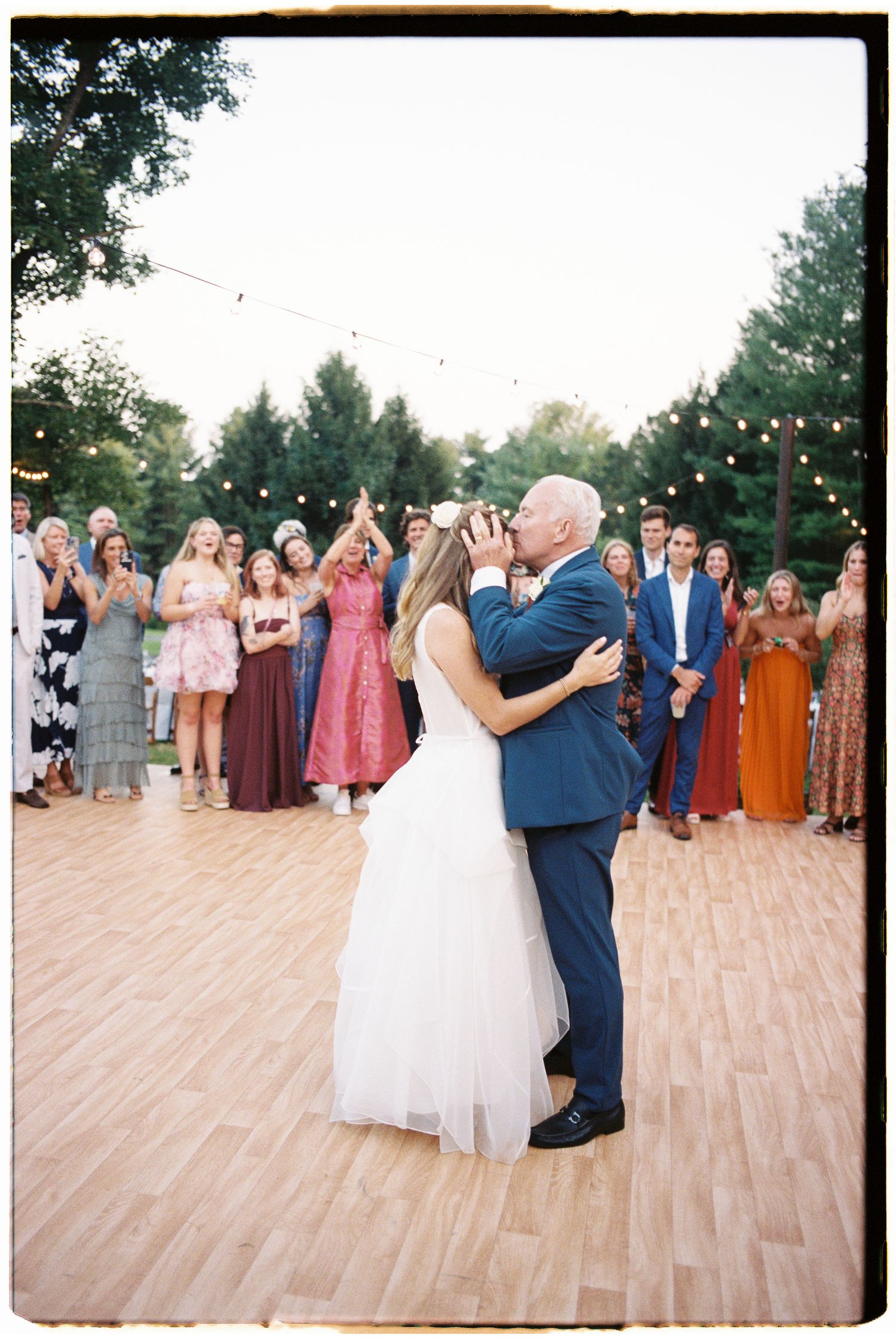Bride and groom share a first dance kiss surrounded by cheering guests at an outdoor wedding reception.