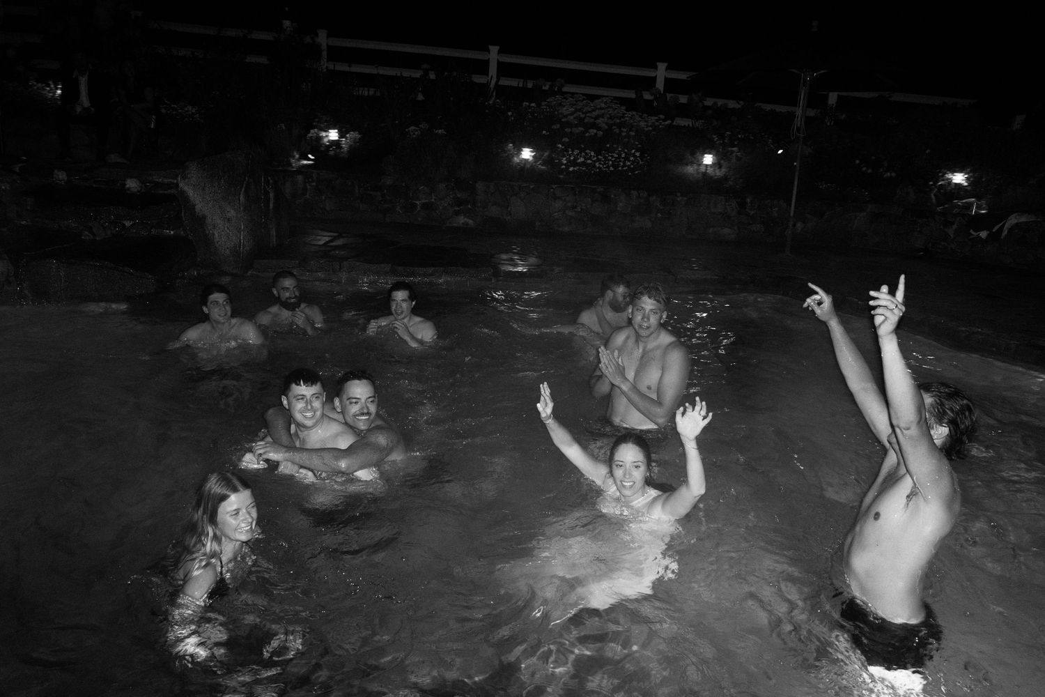 Wedding guests celebrating and swimming in pool at night during outdoor wedding reception party.