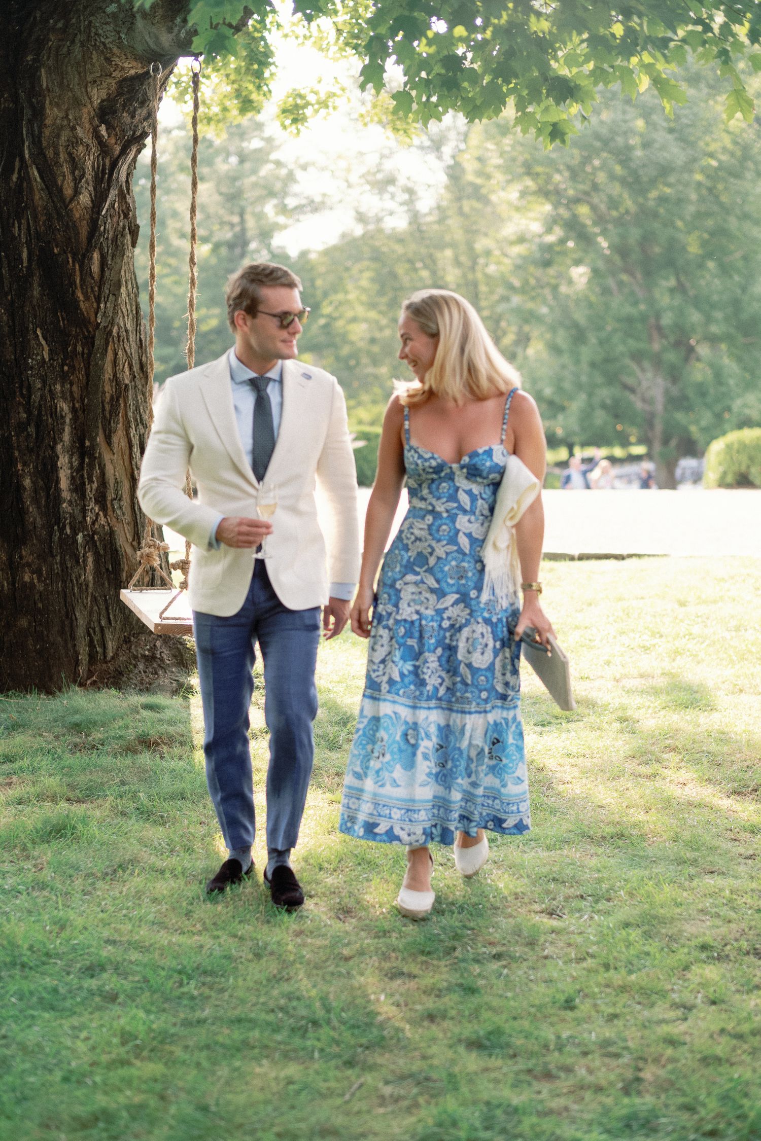 Elegant couple walking hand in hand through a sunny park, man in white blazer, woman in blue floral dress.