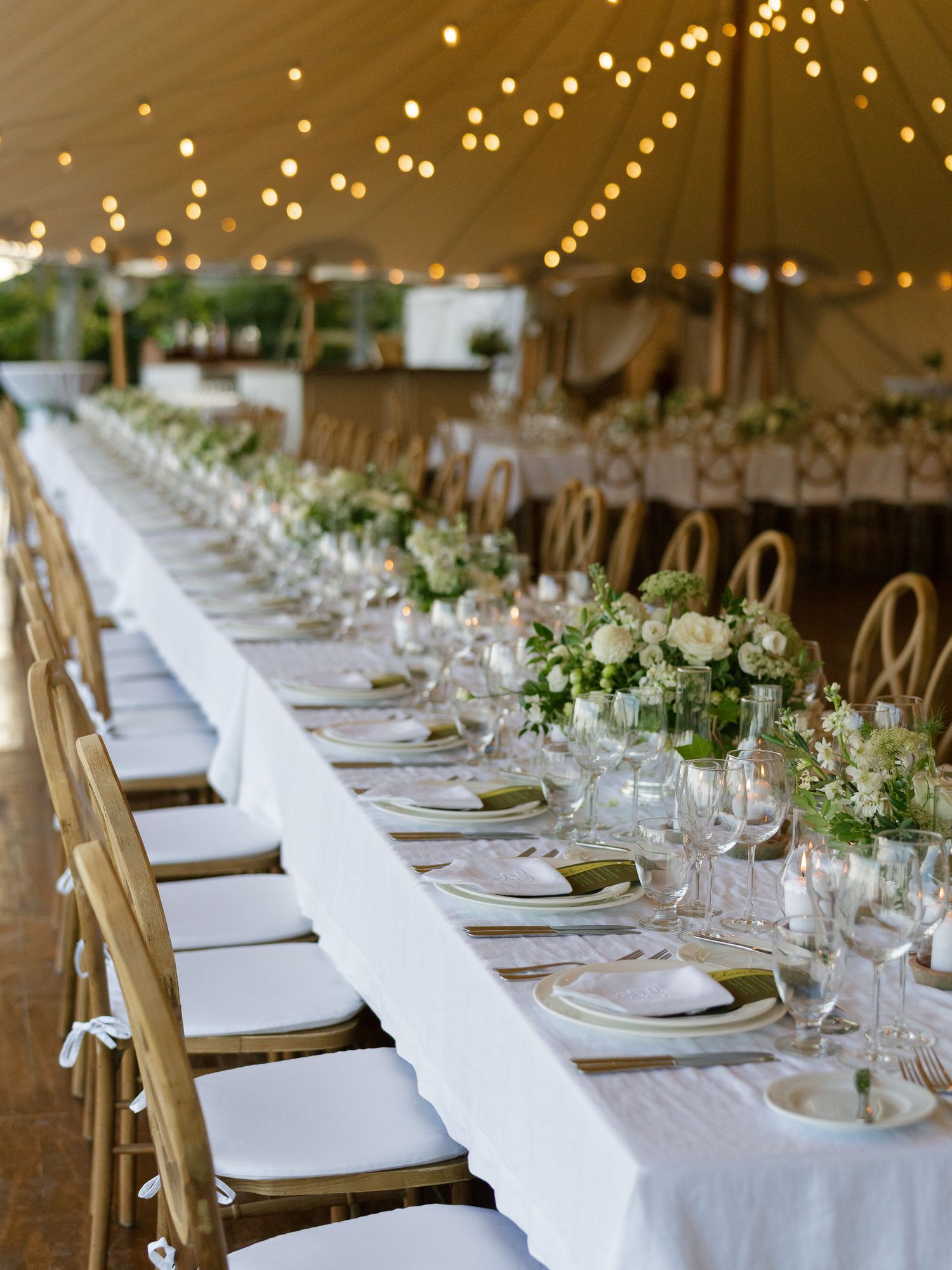 Long wedding reception tables under tent with string lights and white floral centerpieces.