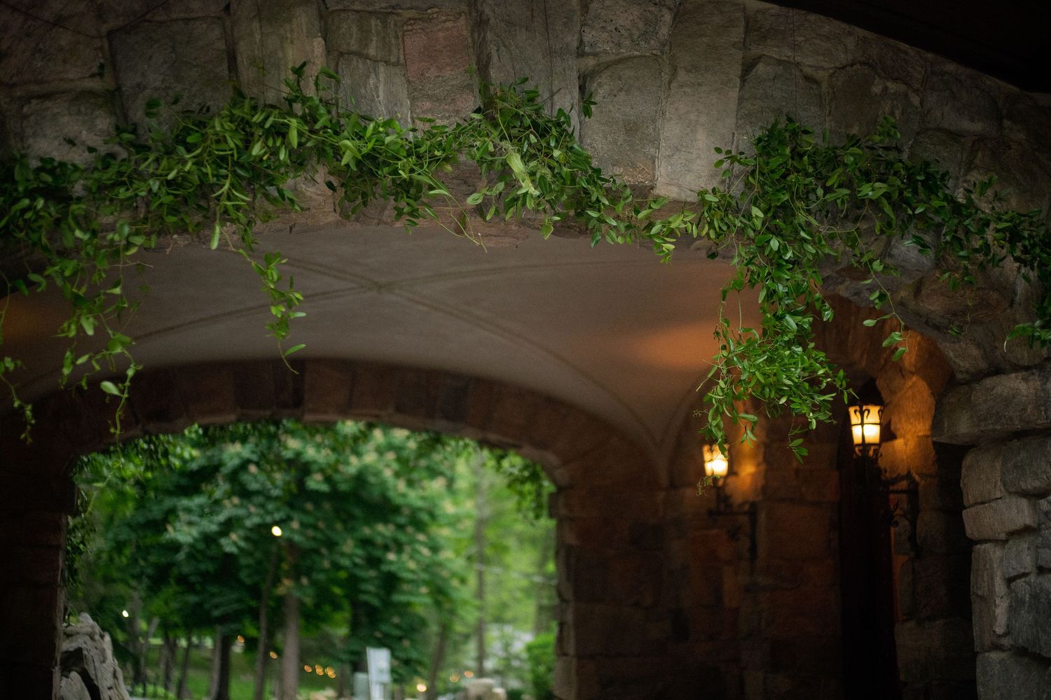Stone archway decorated with lush green garland and warm glowing lanterns at an outdoor wedding venue.