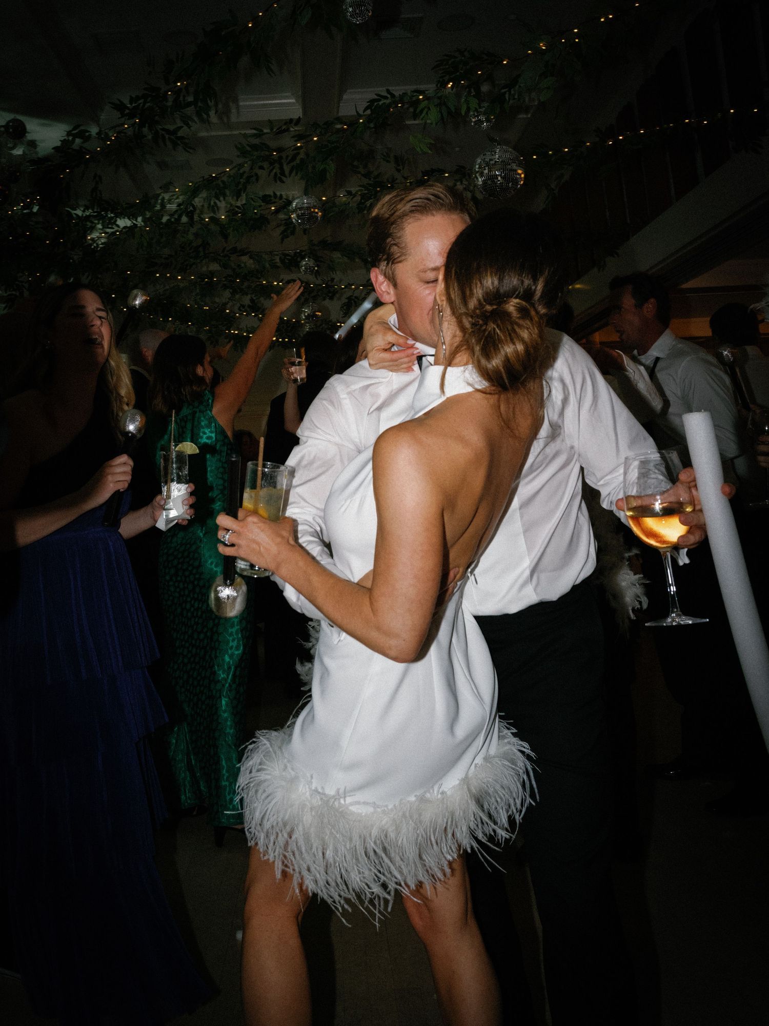 Bride in white feathered mini dress kissing groom on dance floor at lively wedding reception.