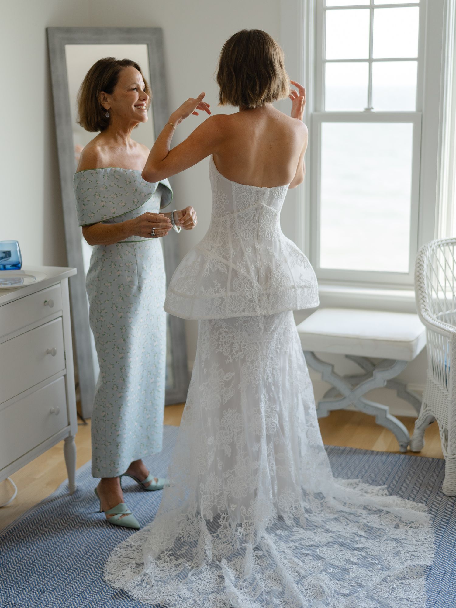 Mother helping bride in strapless lace peplum wedding gown prepare in bright bedroom.