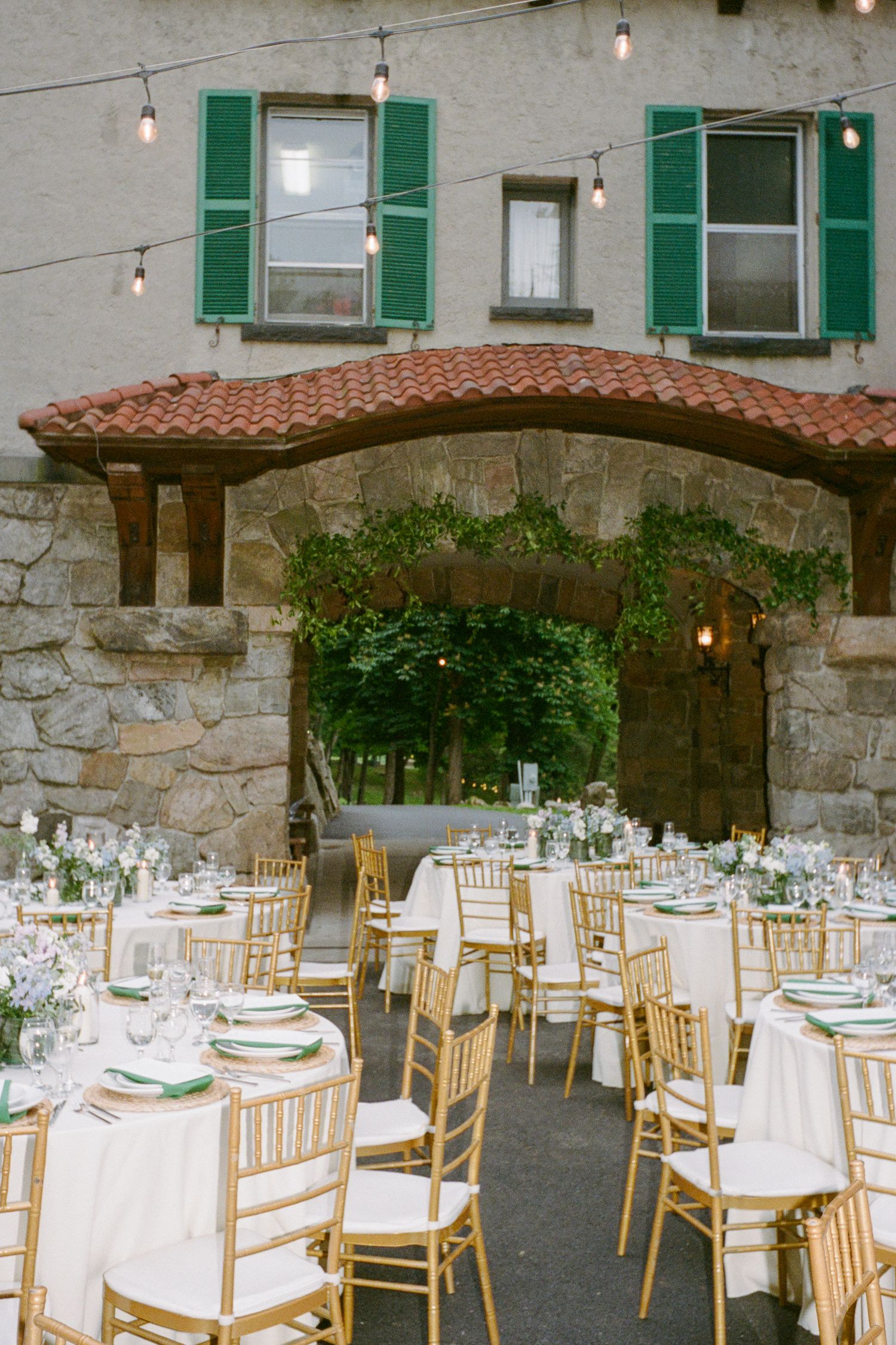 Elegant outdoor wedding reception with gold chiavari chairs, white linens, and string lights at a rustic stone venue.