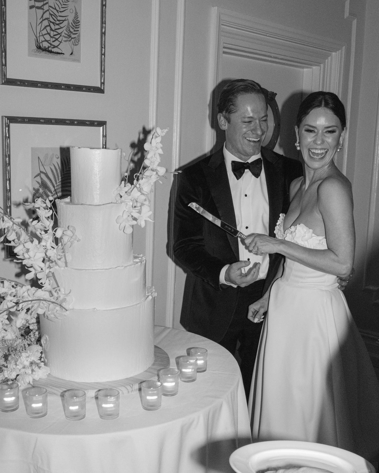 Bride and groom smiling while cutting four-tier white wedding cake at reception.